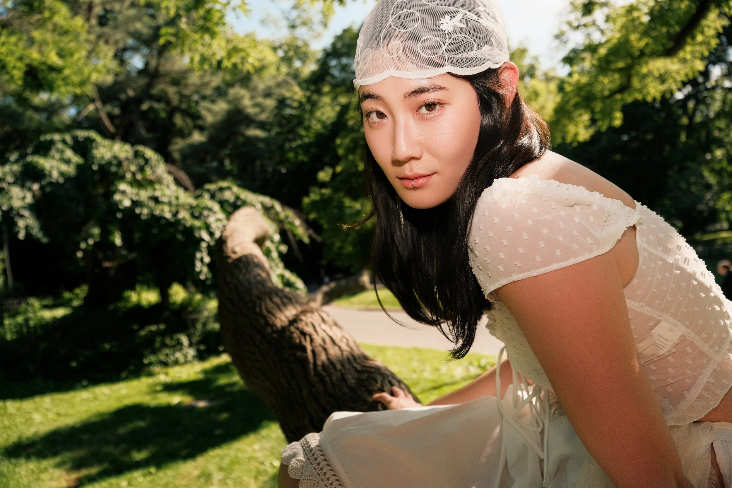 Young woman in a white dress and lace cap outdoors on a sunny day, leaning over a tree trunk in a park.