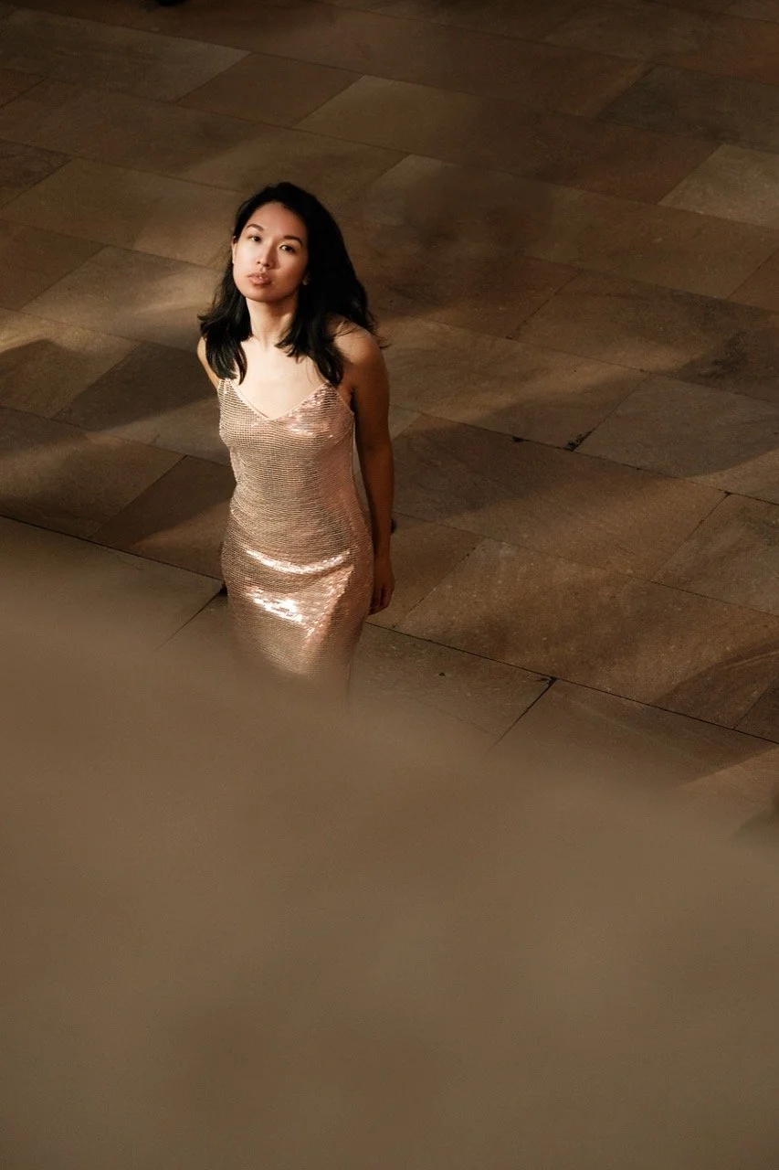 A woman in a shiny, rose-colored dress standing on a tiled floor, looking up at the camera with a neutral expression.