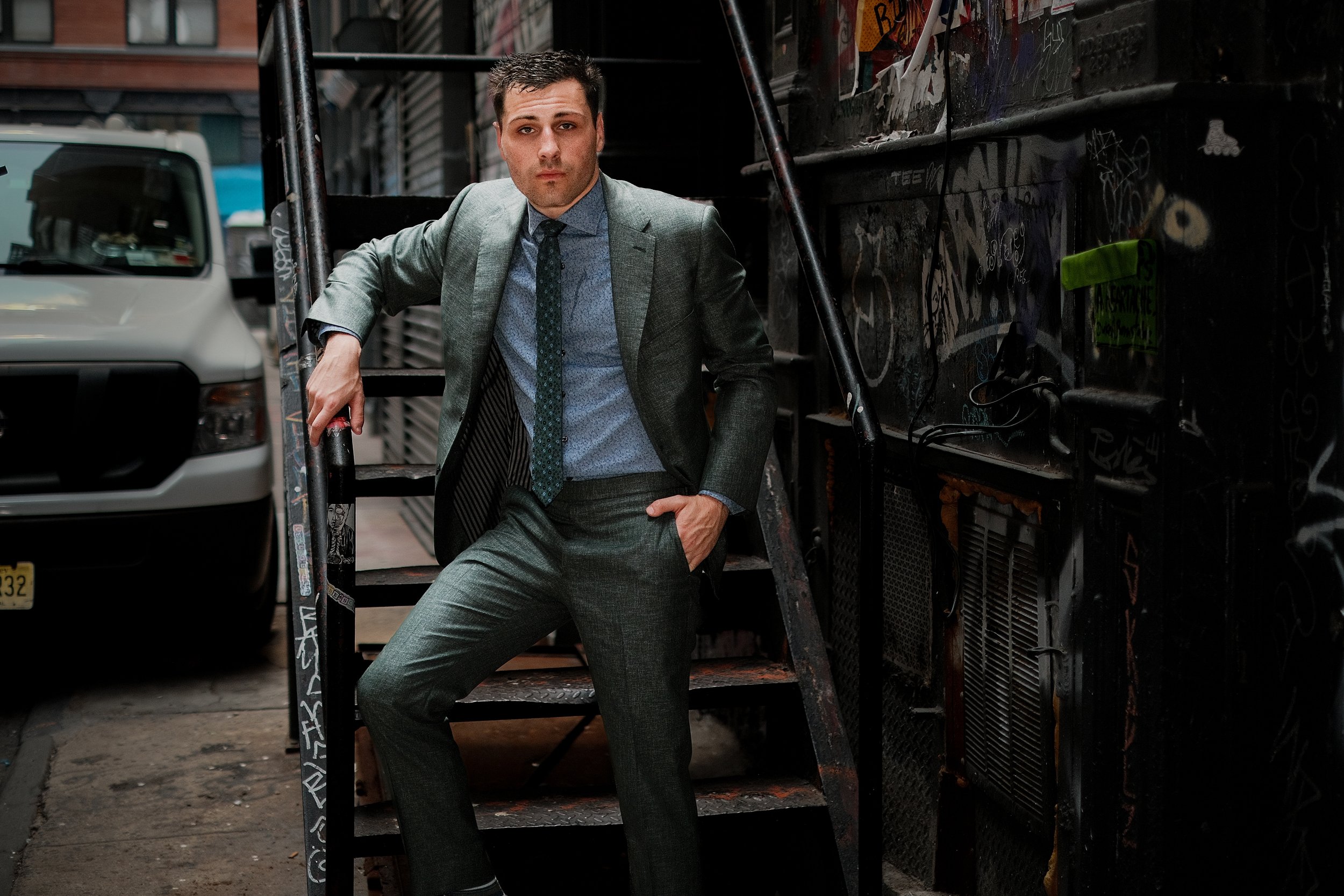 A man wearing a gray suit and tie is leaning against a metal railing in an urban alleyway, with graffiti-covered walls and parked vehicles visible in the background.