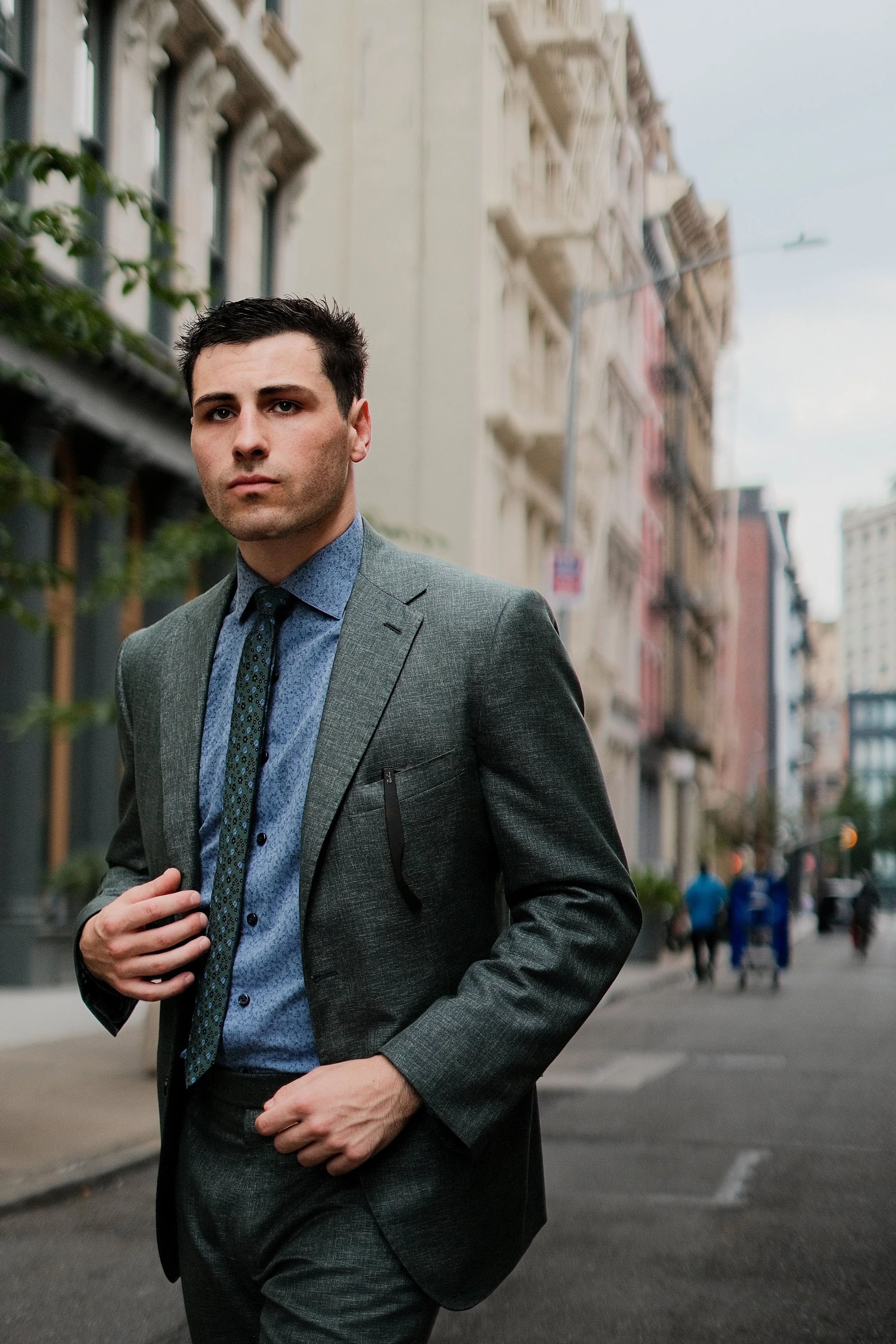 A man in a gray suit and blue shirt walking on a city street with high-rise buildings in the background.
