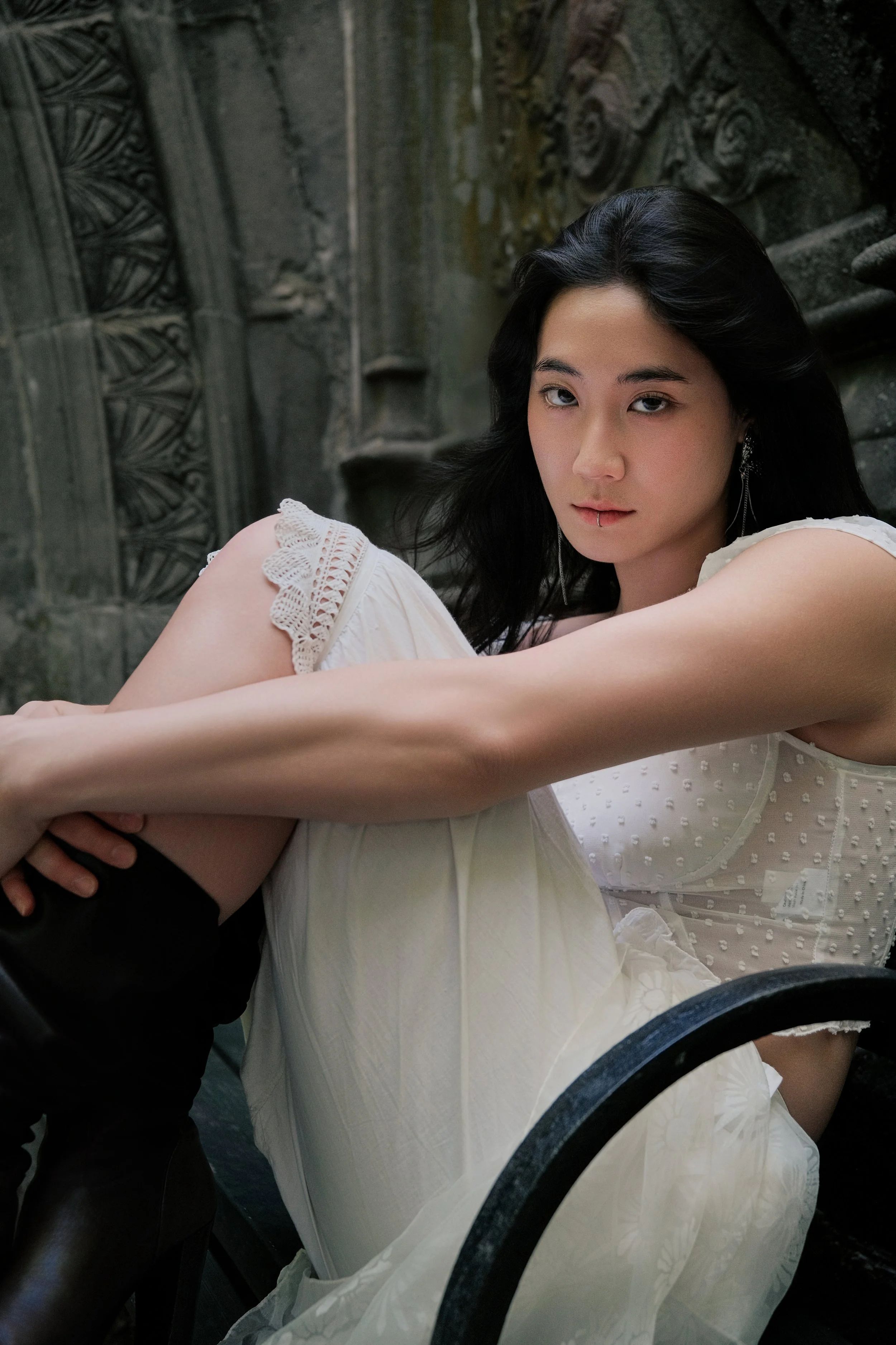 A young woman with black hair, wearing a white dress with lace details and earrings, sitting with her arms around her knees, looking at the camera with a serious expression. The background features an ornate stone or concrete wall with detailed carvi