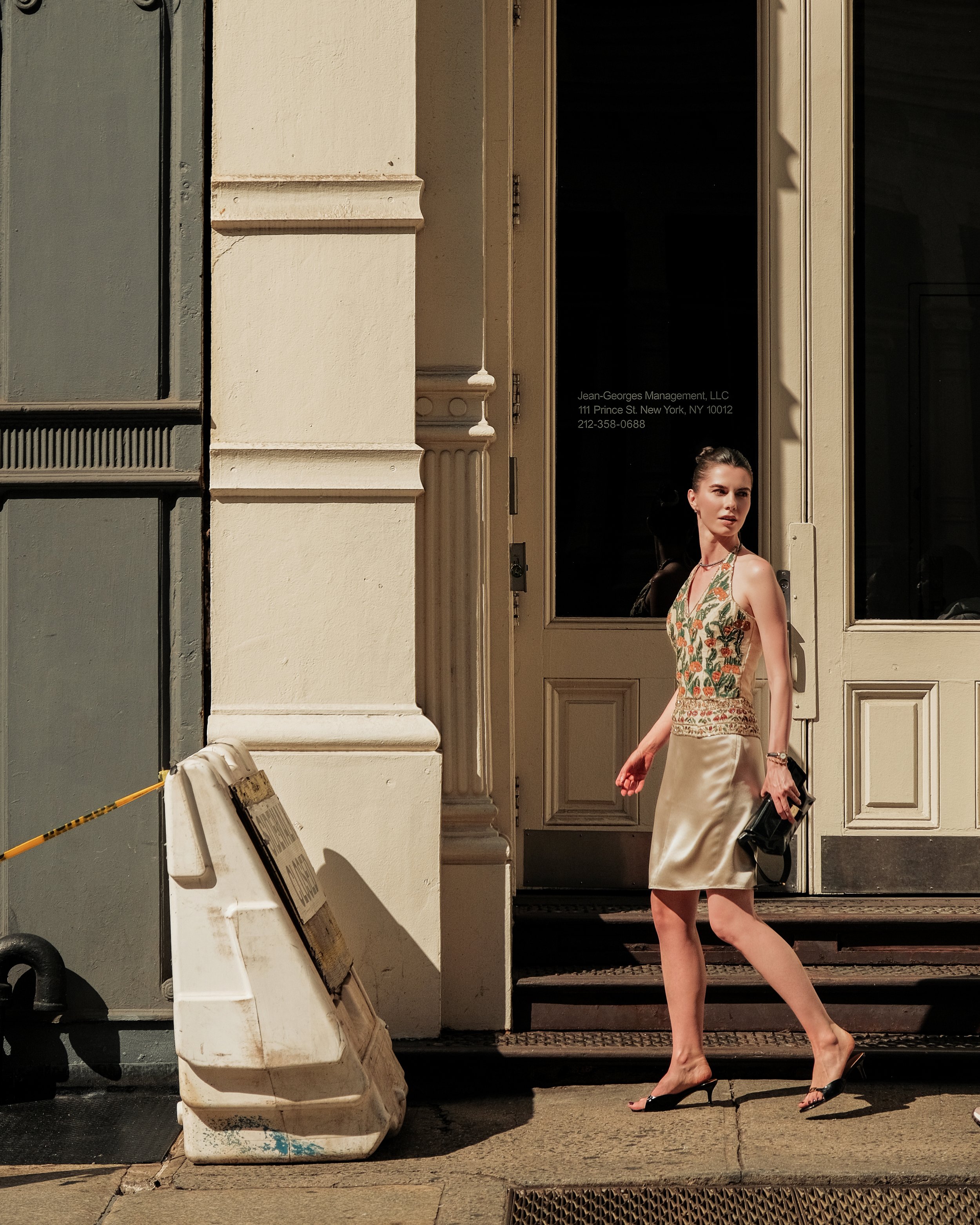 Woman walking past a building with a glass door, wearing a floral dress and black high heel shoes, holding a black handbag.