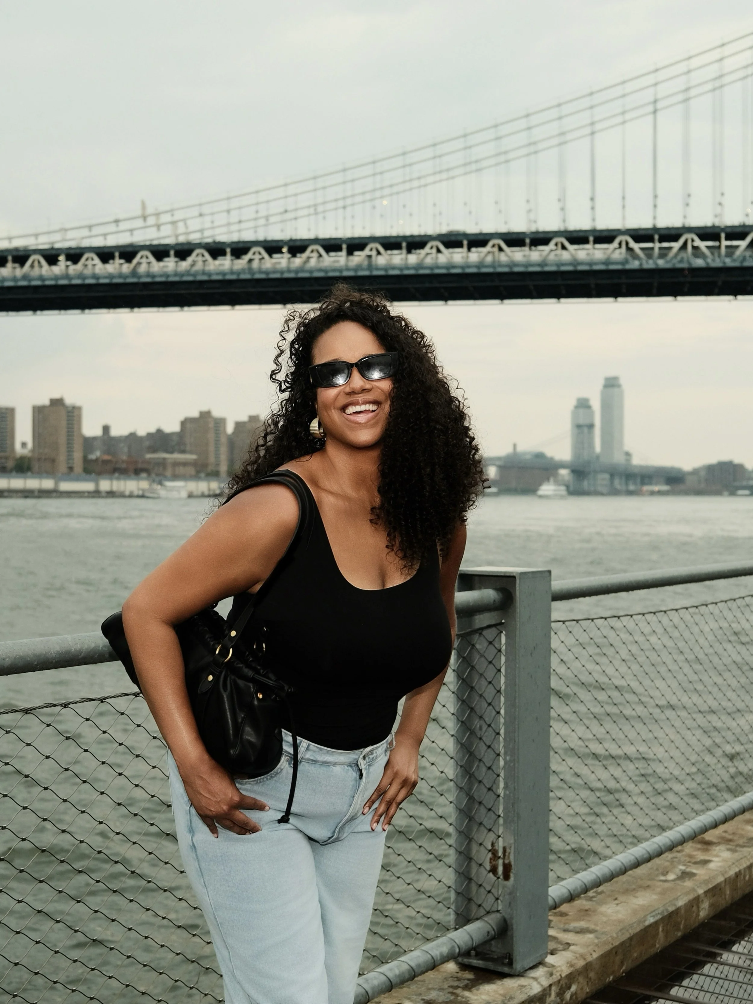 A woman with curly hair wearing sunglasses, a black tank top, and light jeans stands smiling by a waterfront with a bridge and city skyline in the background.