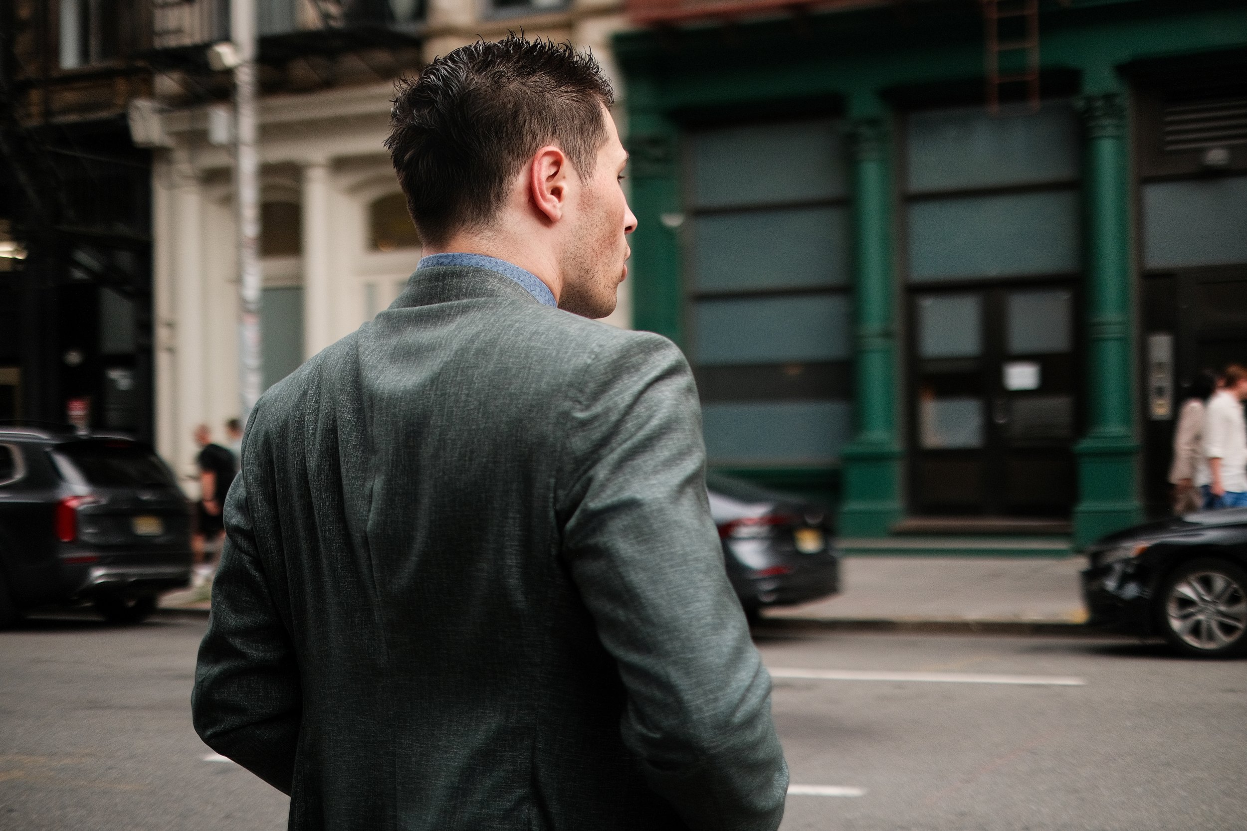 Side profile of a young man with dark, styled hair wearing a gray blazer, walking on the street with cars parked and a building with green columns in the background.