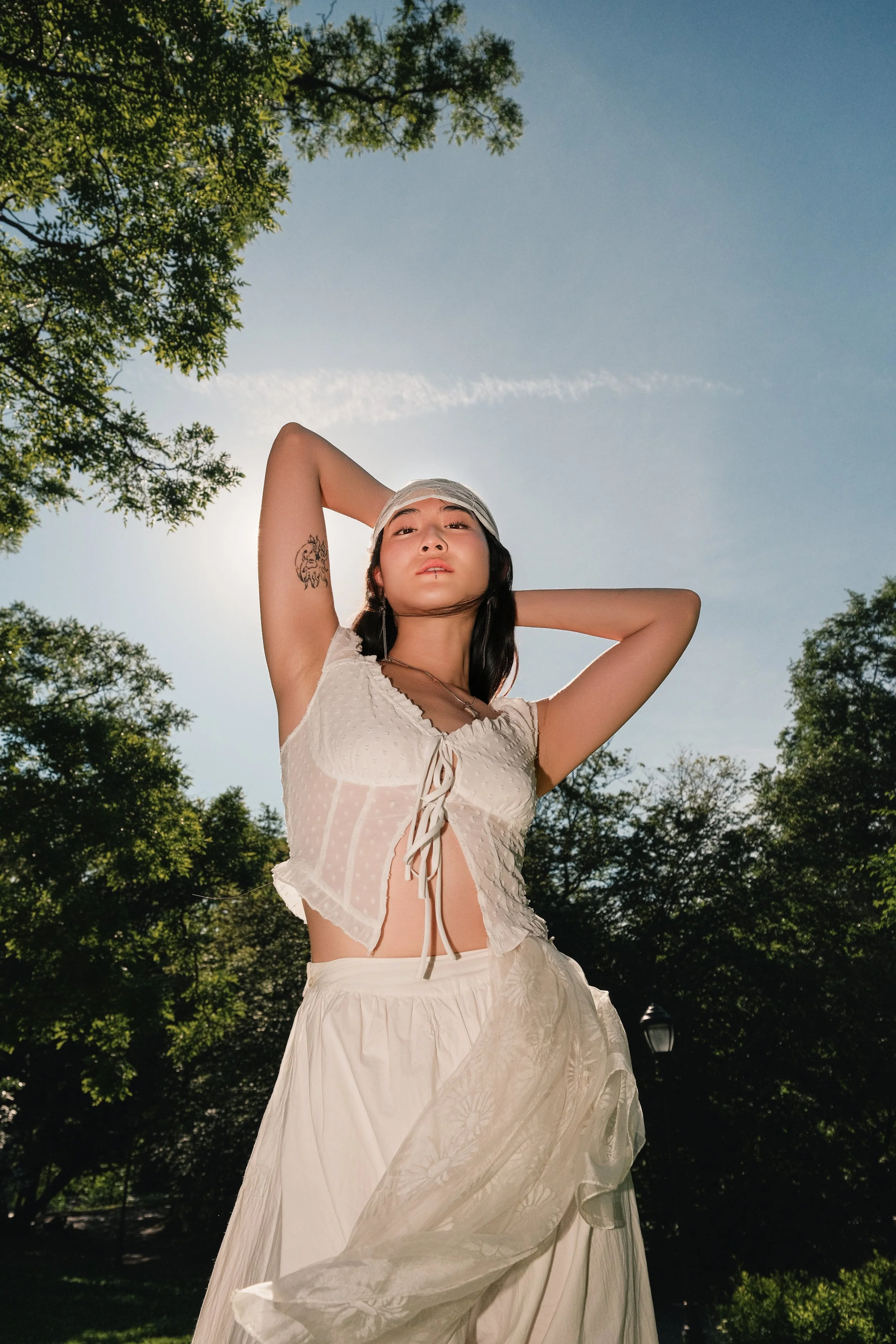 young woman wearing a beige hat and white outfit standing outdoors with trees and a clear blue sky in the background