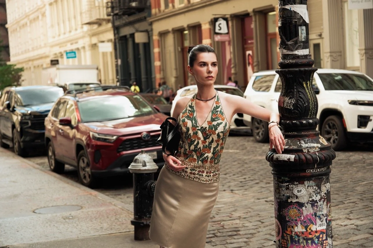 A woman poses on a city street near a black pole covered in stickers, with parked cars and historic buildings in the background.