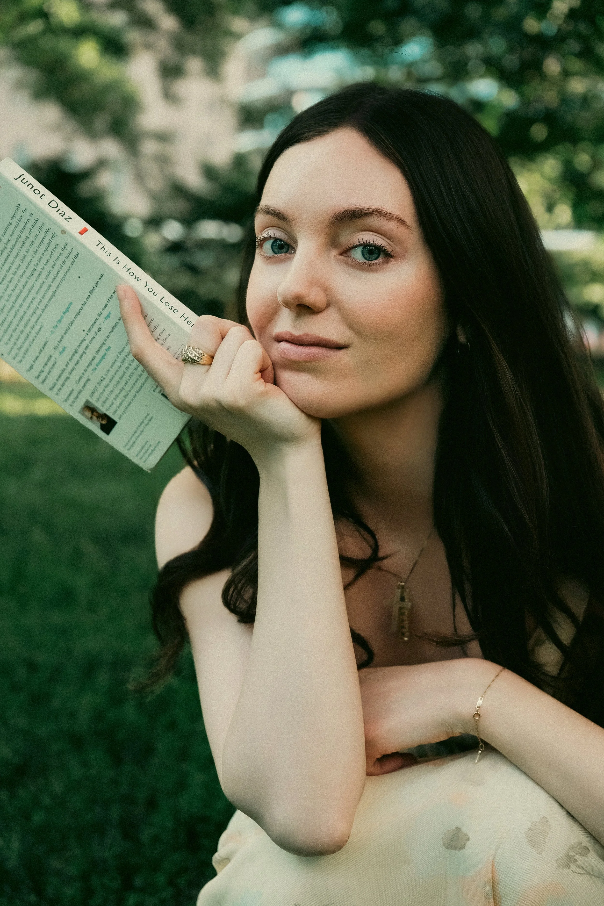 A young woman with long dark hair and blue eyes sitting outdoors on grass, holding a newspaper close to her face. She is wearing a beige top and has jewelry including rings, a necklace, and a bracelet.