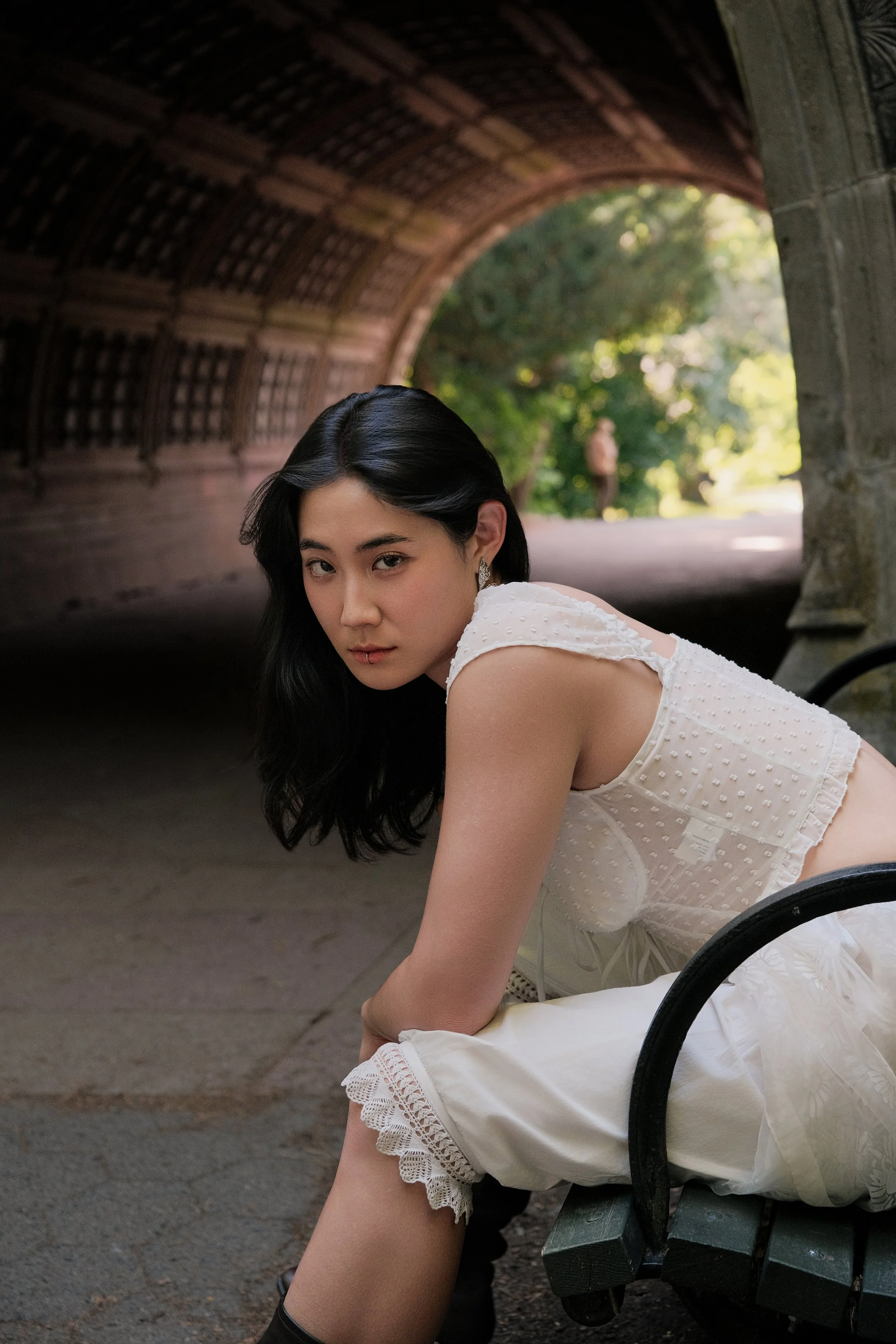 A young woman with black hair, wearing a white dress with lace details, sitting on a park bench under a wooden arch structure. She is looking at the camera with a serious expression, and there is a background of green trees and sunlight filtering thr
