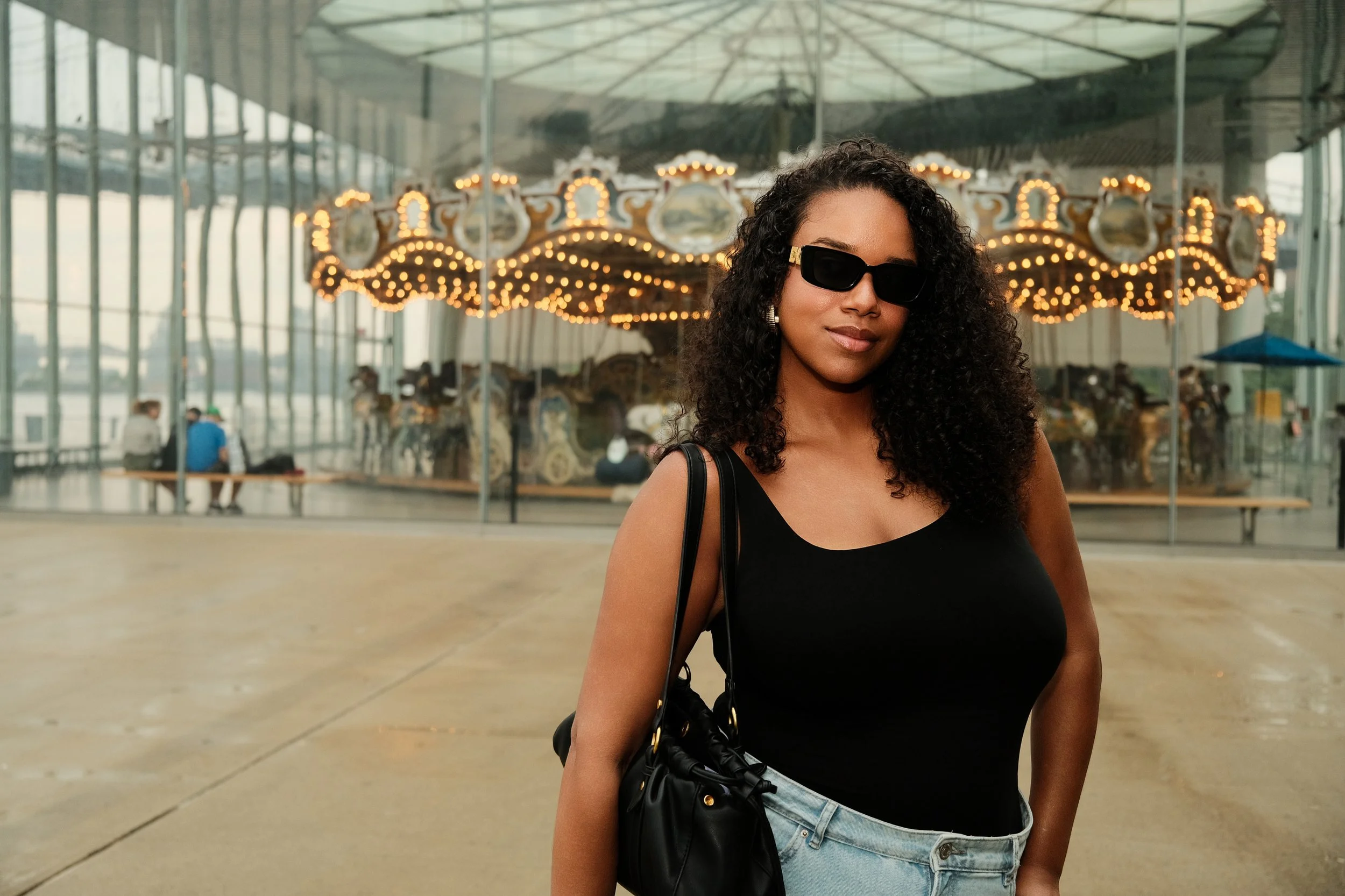 A woman with curly hair wearing sunglasses and a black top, standing in front of a carousel at an amusement park or fairground.