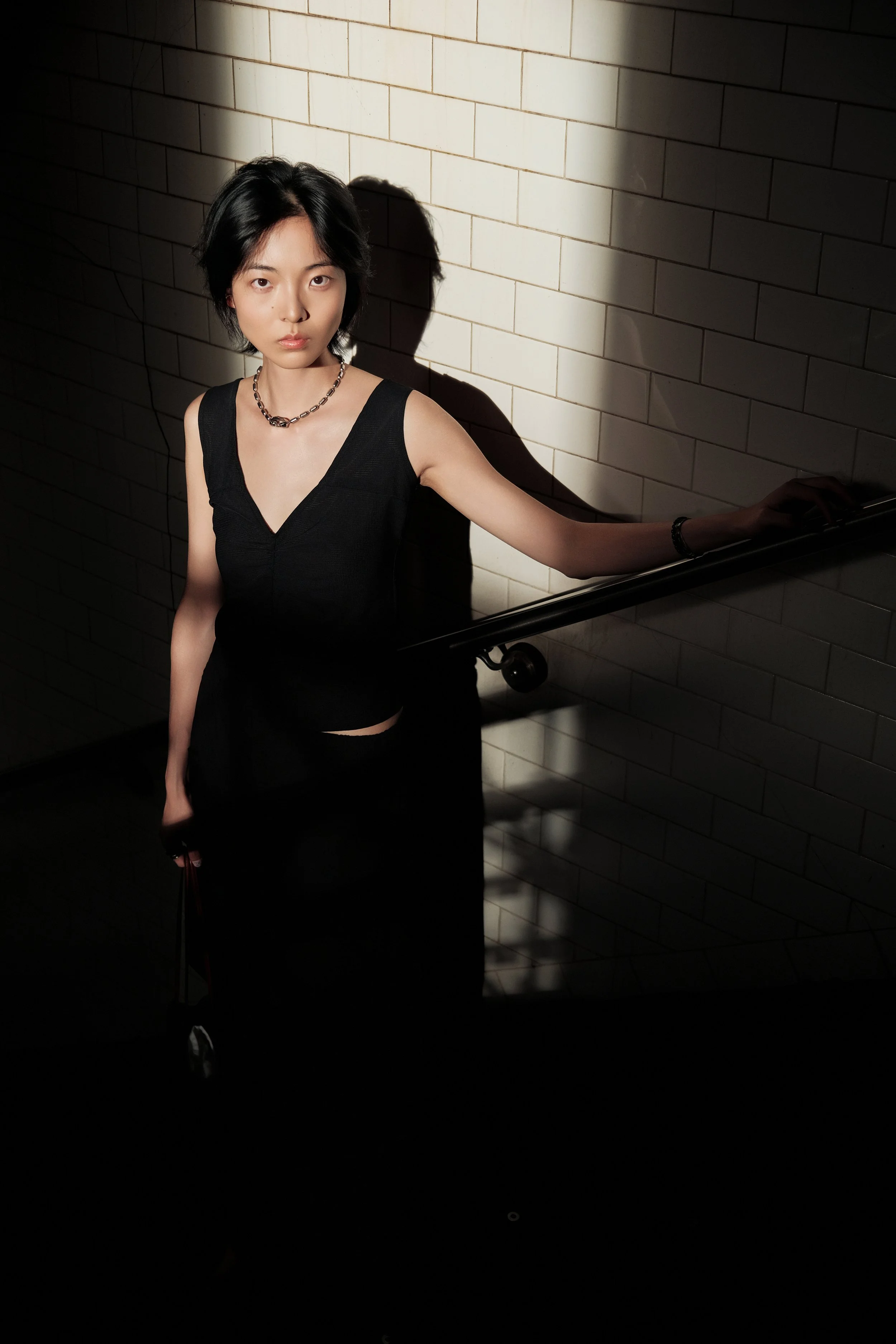 A young woman with short black hair wearing a black sleeveless top, a chain necklace, and a bracelet, standing against a white tiled wall. She is holding onto a handrail with her right hand, and a shadow of her figure is cast on the wall behind her. 