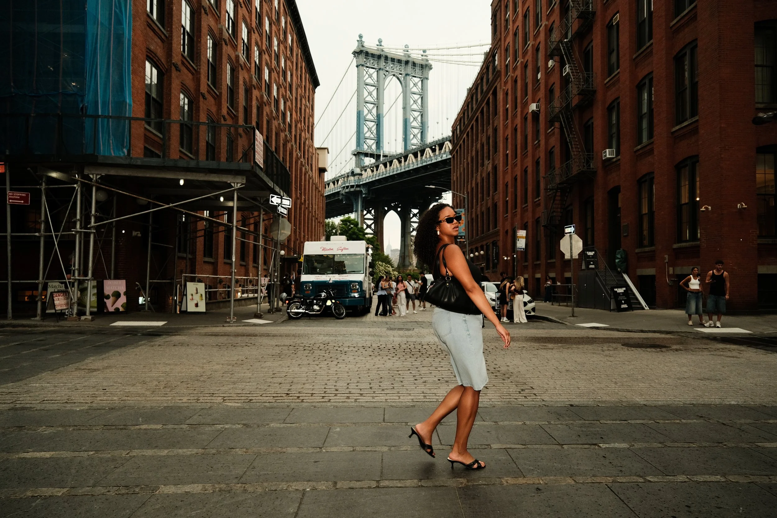 A woman with curly hair wearing sunglasses, a black sleeveless top, light jeans, and black high heels walking across a city street with brick buildings and a bridge in the background.