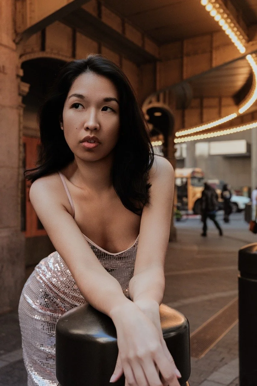 A young woman with black hair wearing a sparkly, light-colored dress leaning on a barrier in an urban setting, with lights and people in the background.