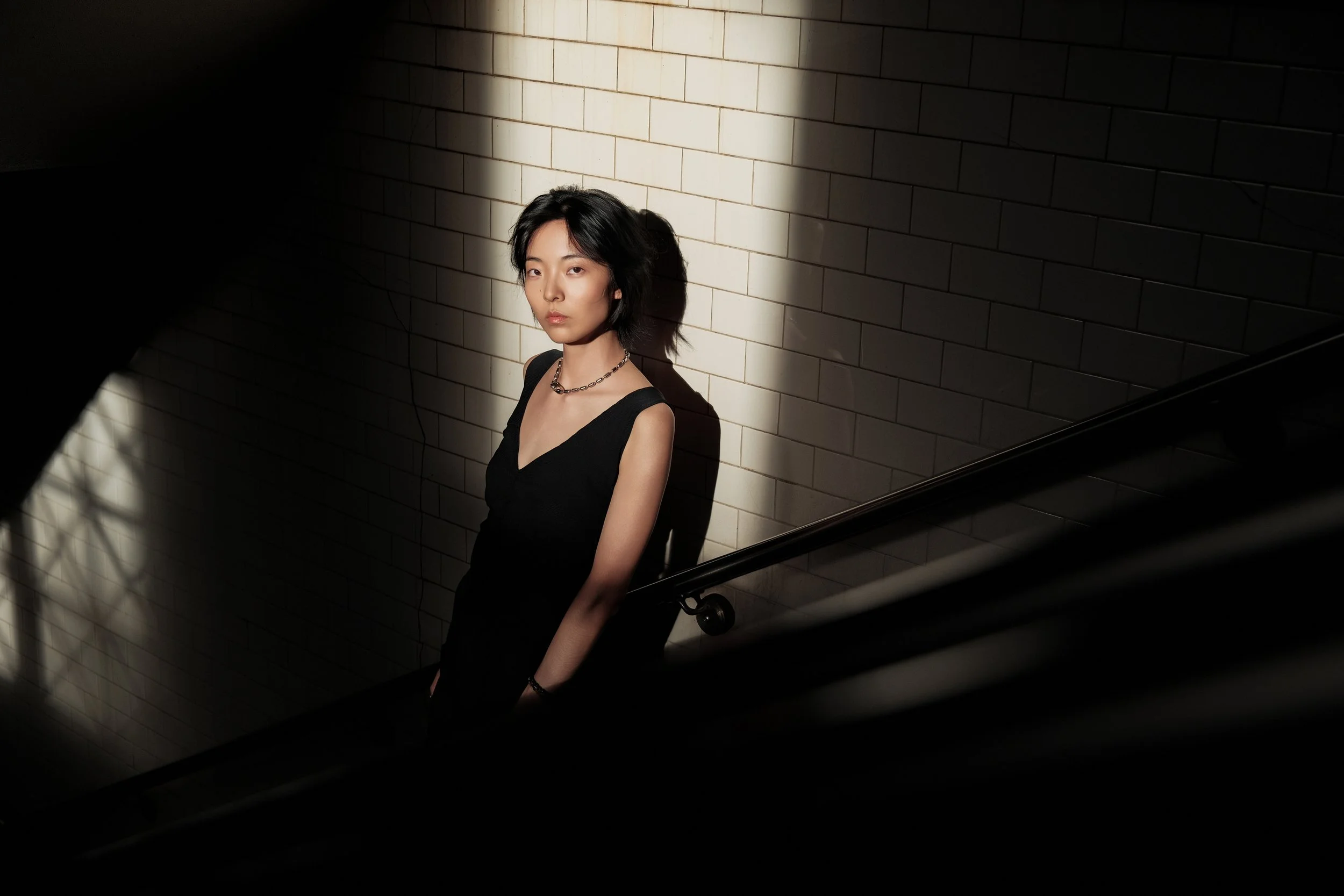 A young woman with short black hair wearing a black sleeveless top and a chain necklace, standing against a white tiled wall in a dimly lit setting with sunlight casting shadows.