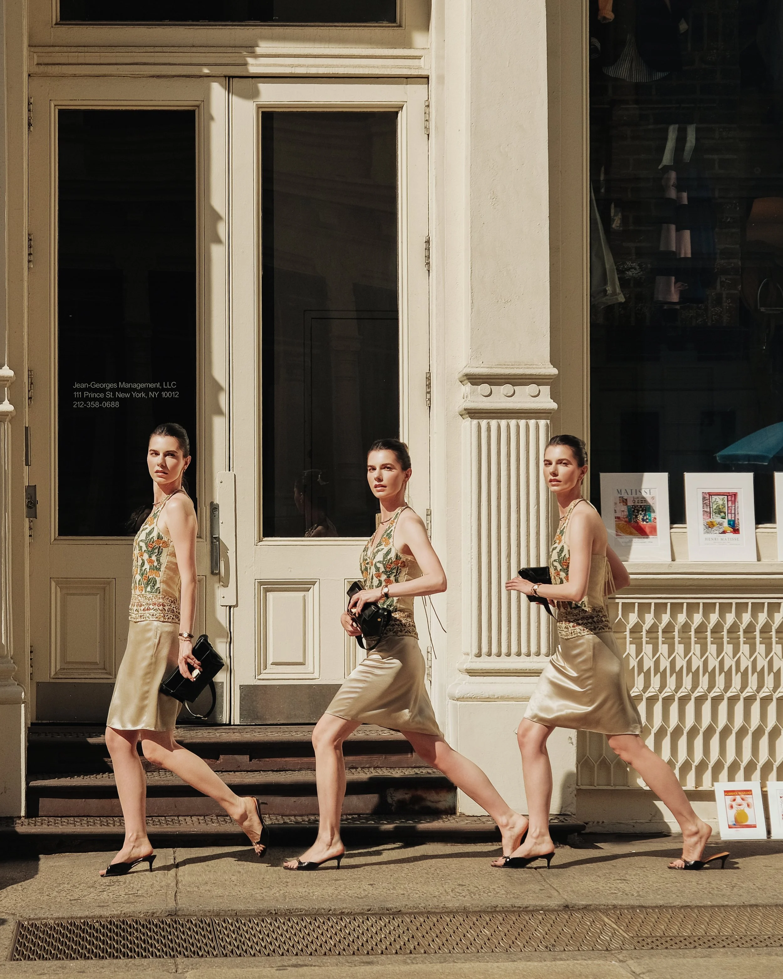 A street scene showing a storefront with four women dressed in matching beige and floral outfits walking across the sidewalk in front of the store, which has large glass windows and a white door. The women appear to be walking in different poses, wit