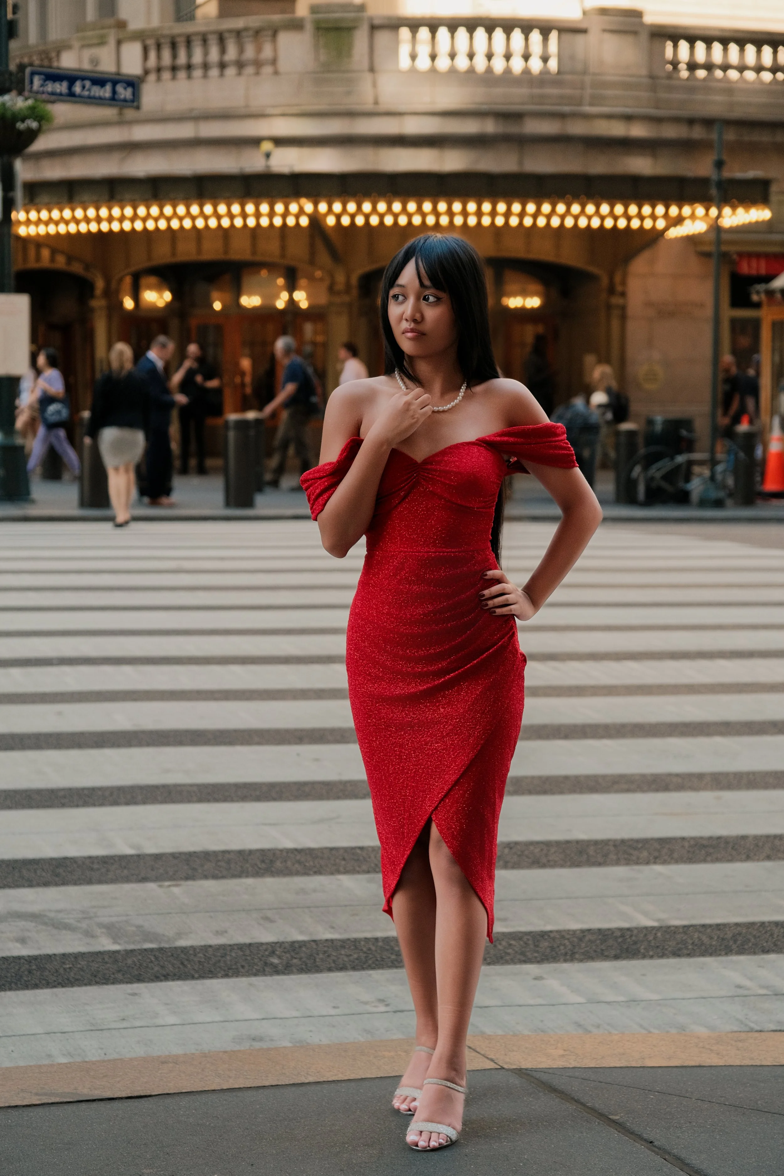 A young woman in a red off-shoulder gown stands on a crosswalk in front of a building with golden lighting in an urban setting. She is wearing a pearl necklace and beige high-heeled sandals.