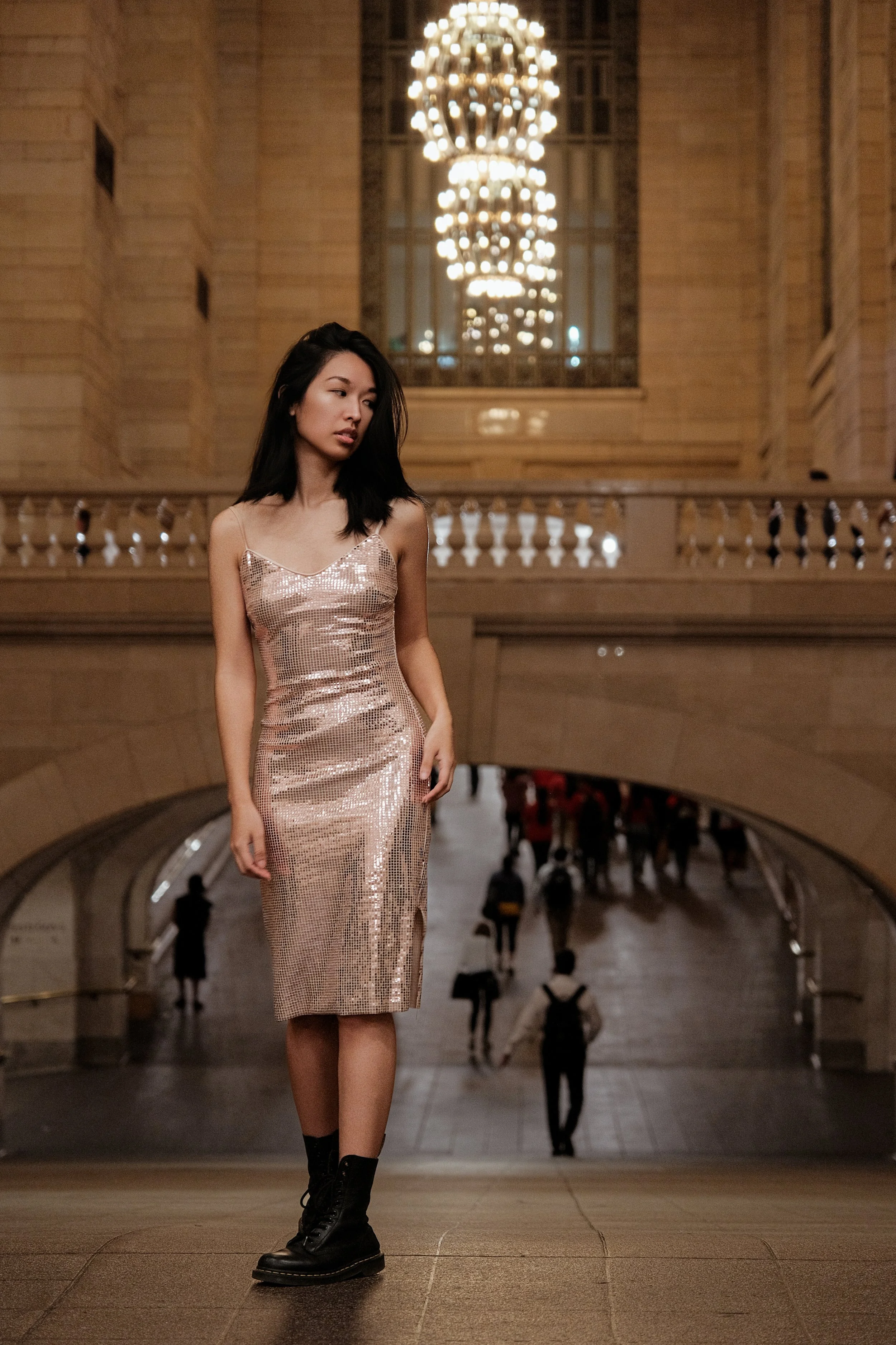 A young woman in a gold sequined dress and black combat boots walking down an escalator in a grand indoor setting with ornate architecture and bright chandeliers.