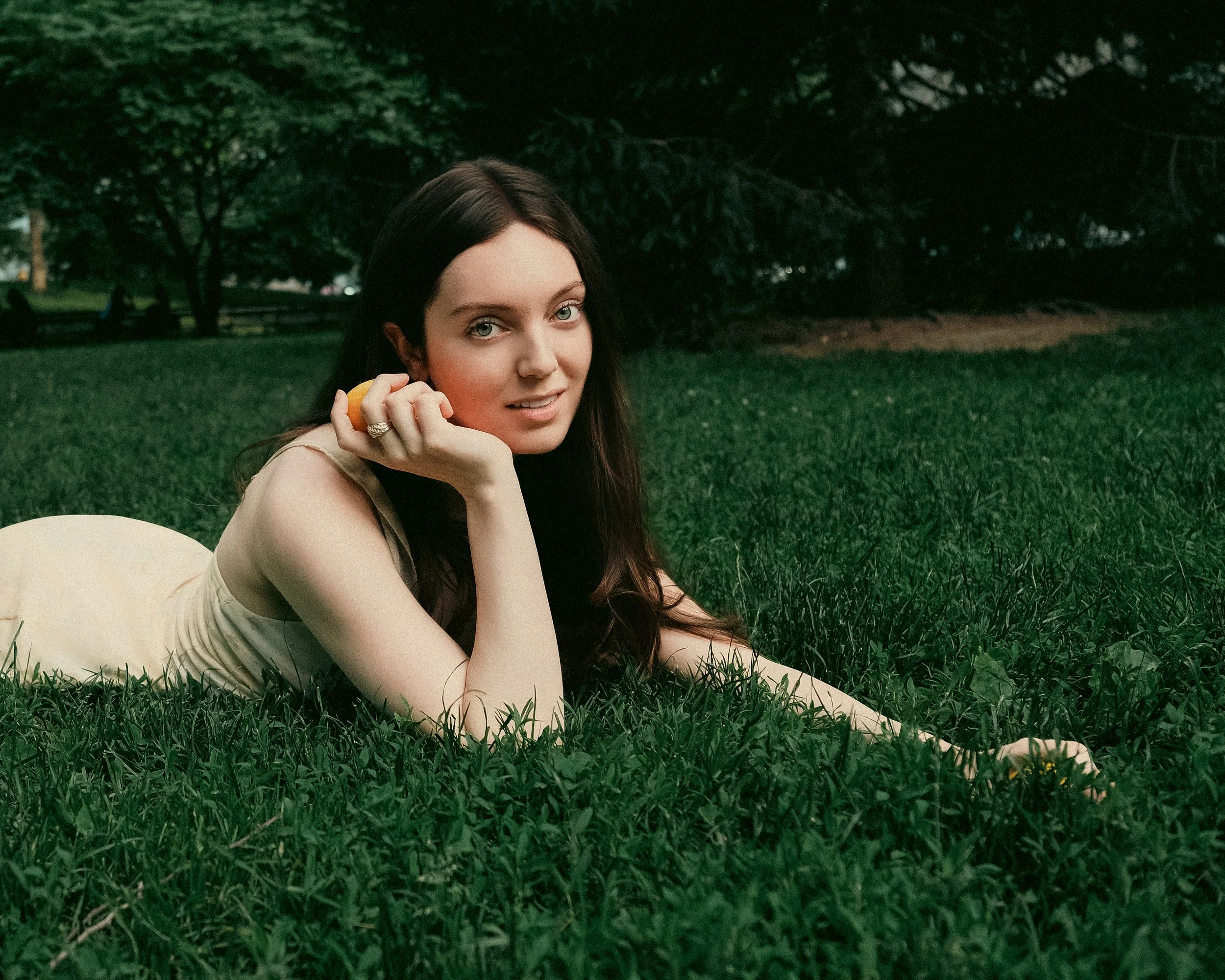 A young woman with long dark hair and blue eyes lying on grass in a park, holding an orange fruit near her face, with trees and a bench in the background.