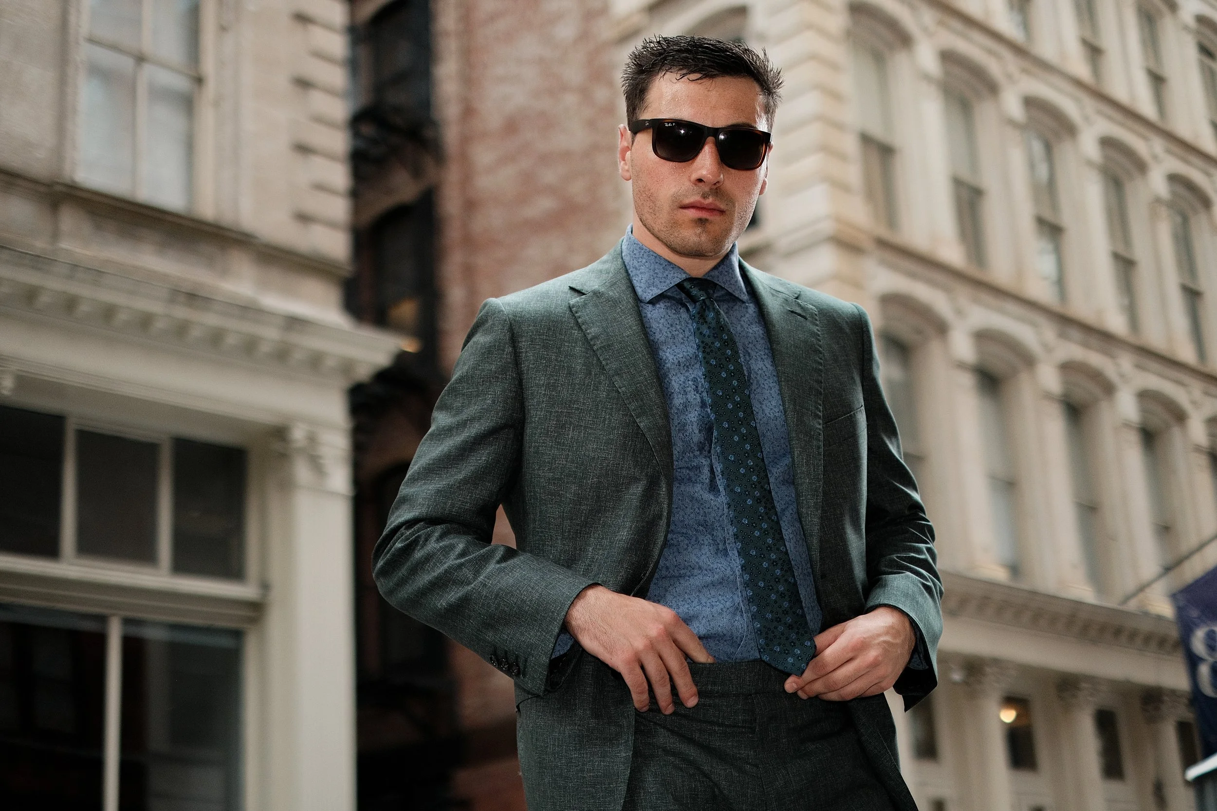 A man wearing a gray suit, blue shirt, and dark tie, with black sunglasses, standing outdoors in front of a city building with ornate architecture.