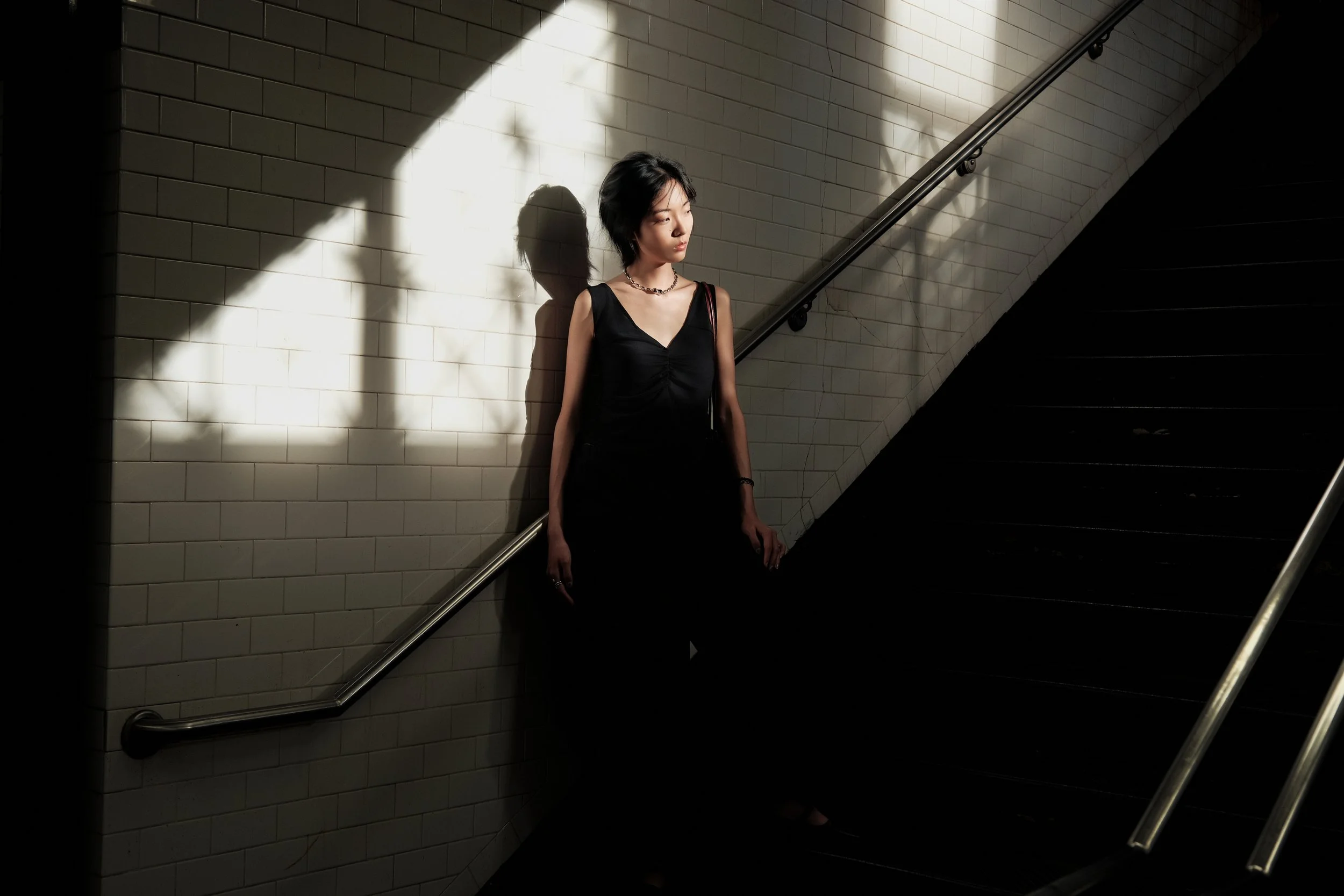 A woman in a black sleeveless dress with a silver necklace stands against a white tiled wall, illuminated by a beam of light, with stairs and a handrail to her right.