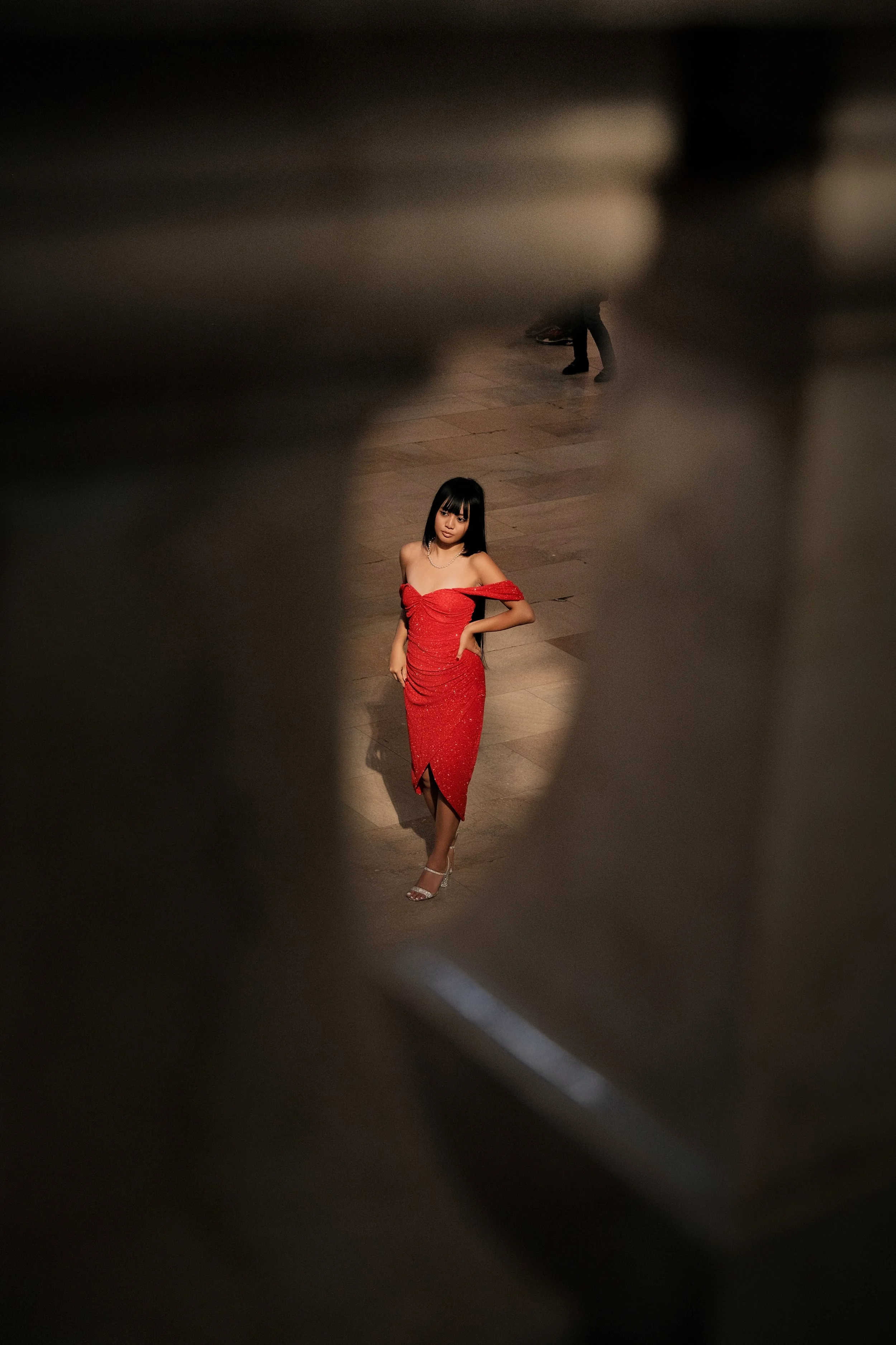 A woman in a red dress standing on a wooden floor, seen through an opening in a blurred black surface.