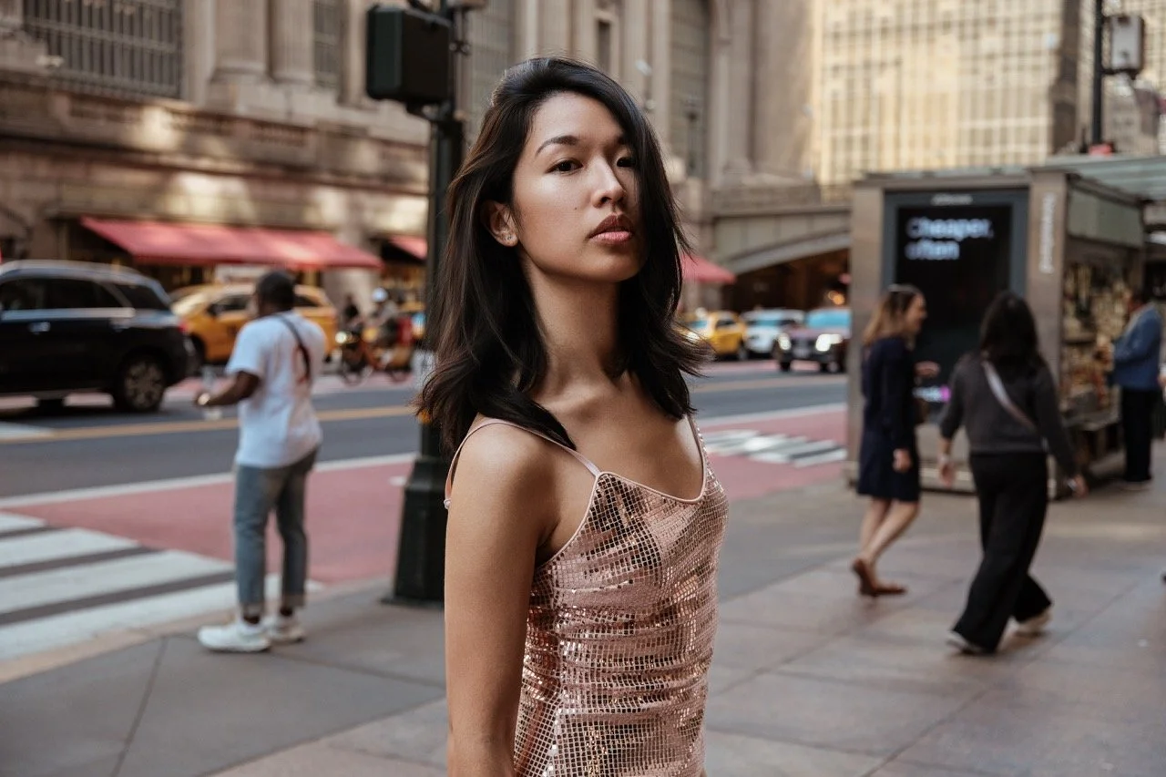 A young woman with dark hair in a rose gold, sequined slip dress standing on a city sidewalk with a crosswalk, taxis, cars, and pedestrians in the background.