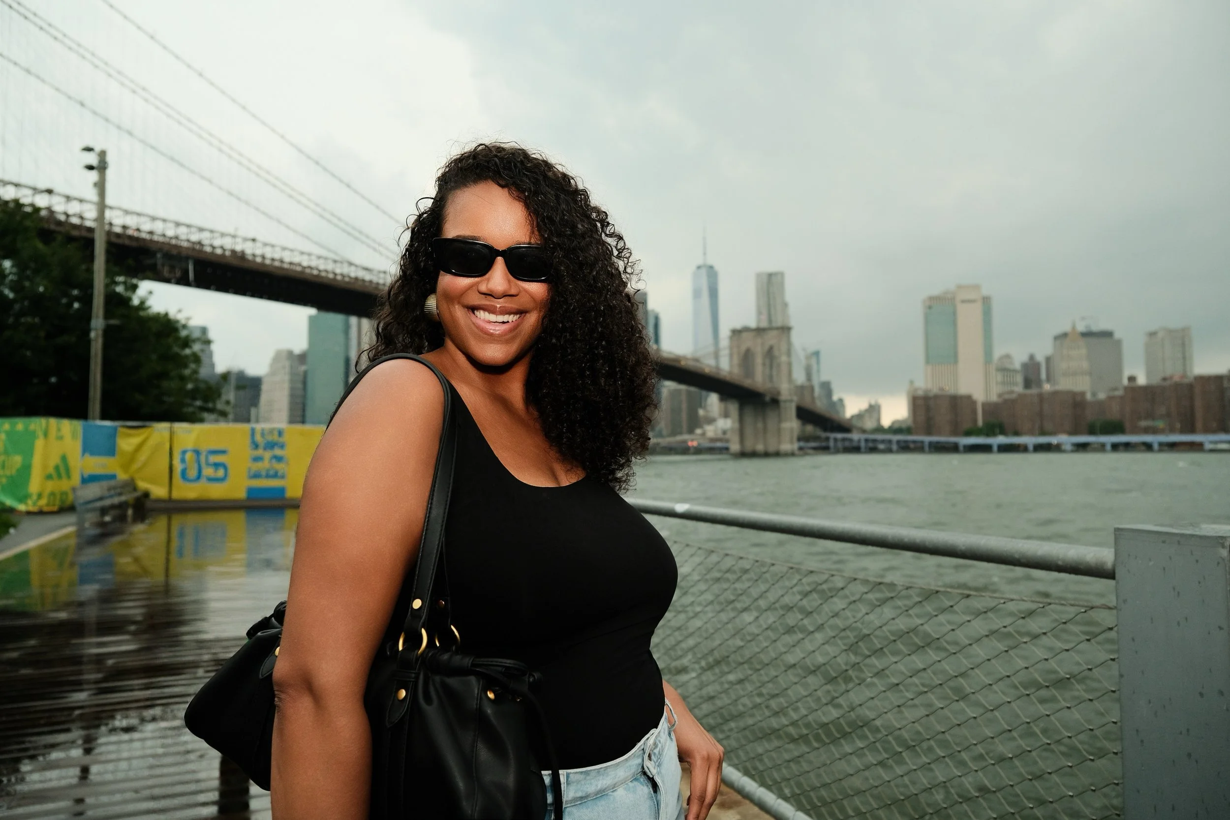Woman with curly hair wearing sunglasses and a black sleeveless top, standing near the water with city skyline and Brooklyn Bridge in the background.
