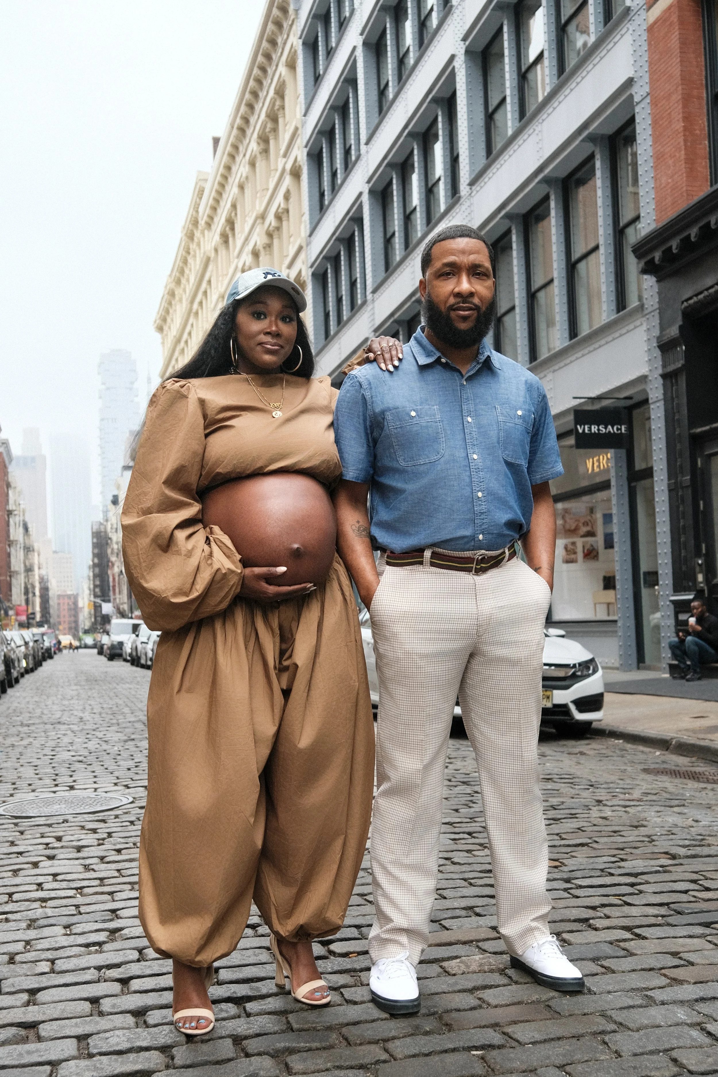 A pregnant woman and a man standing on a cobblestone street in an urban area. The woman is dressed in a tan outfit with a baseball cap, hoop earrings, gold necklace, and high heels. She is cradling her pregnant belly with one hand. The man is wearing