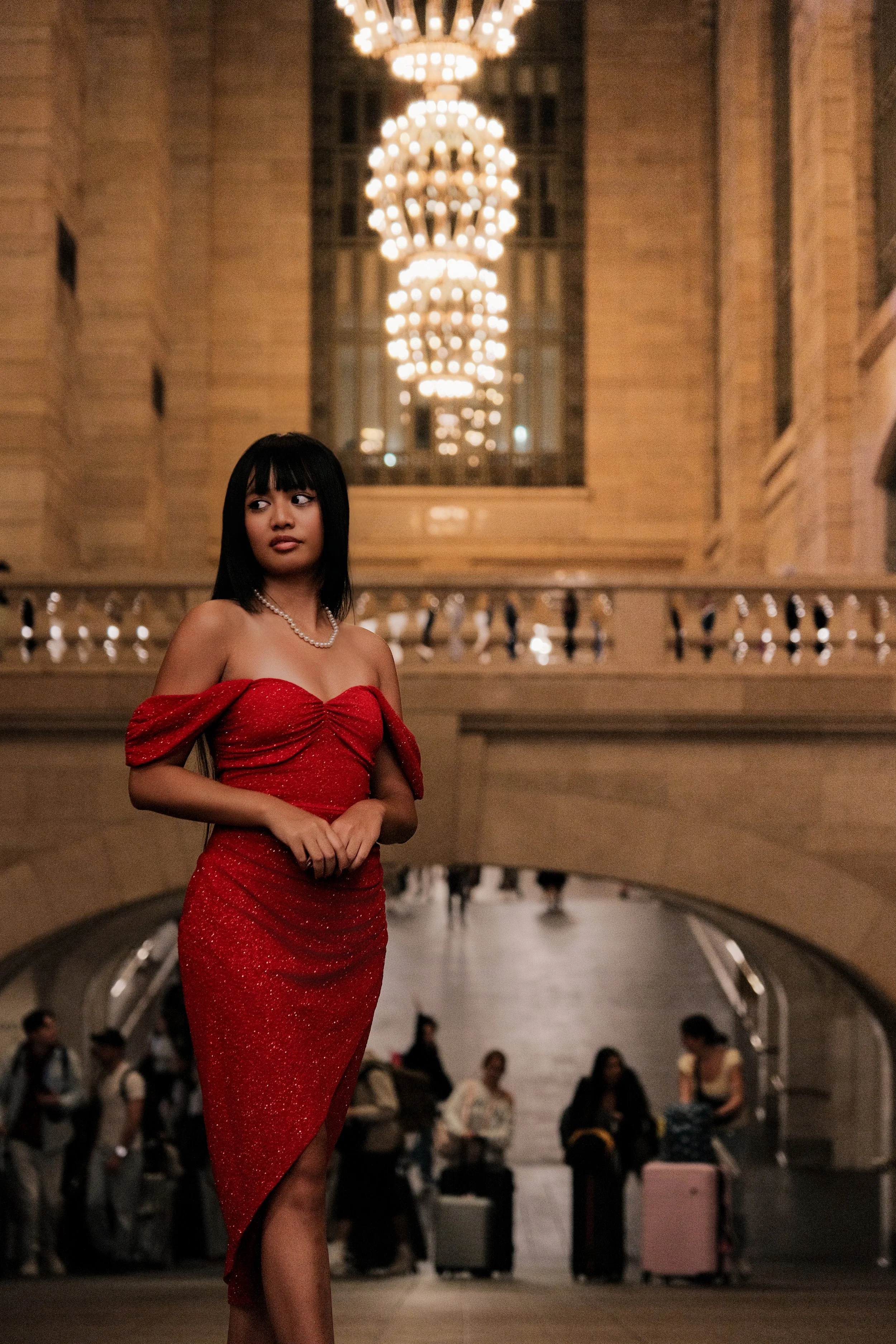 A woman in a red, off-the-shoulder gown with a slit, stands in an elegant, well-lit hallway with a large chandelier overhead. She is wearing a pearl necklace and has shoulder-length dark hair with bangs. People are seen in the background, some with l
