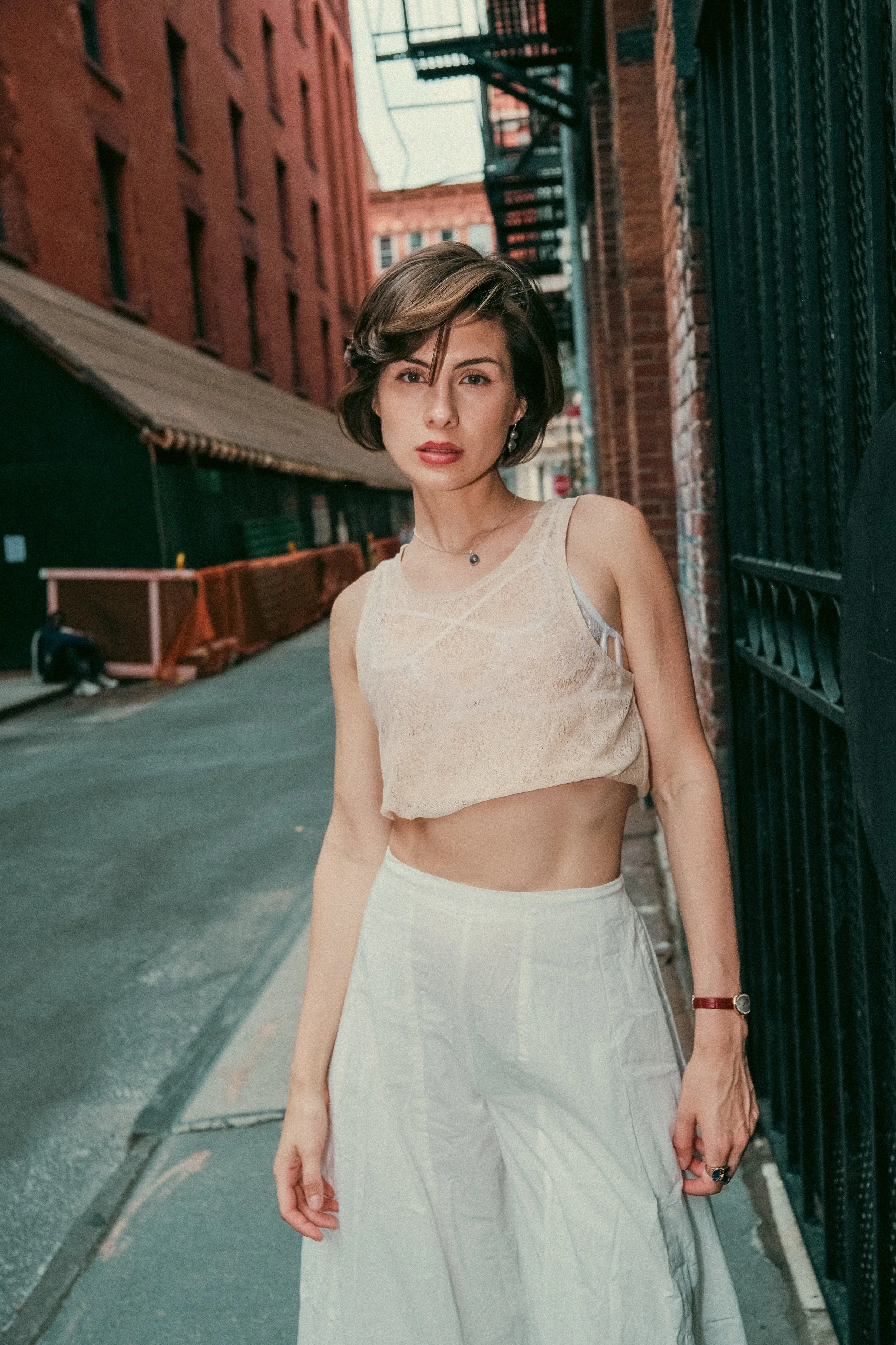 A woman with short brown hair standing on a city sidewalk, wearing a sleeveless cream-colored top and a light-colored skirt, with brick buildings and fire escapes in the background.