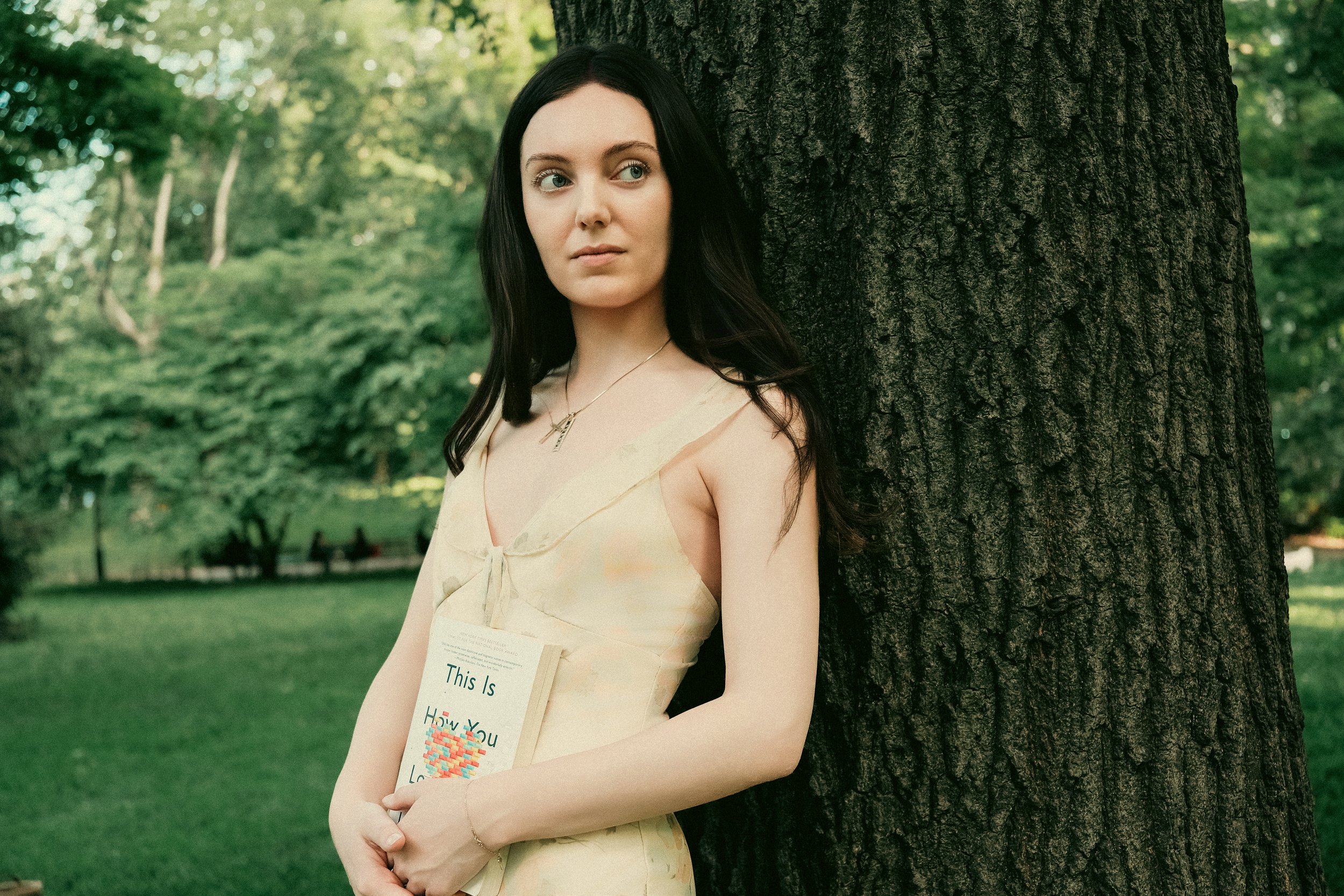A young woman with long dark hair stands next to a tree in a park, holding a piece of paper with colorful text and an illustration. She is wearing a light-colored dress and looking to the side with a neutral expression.