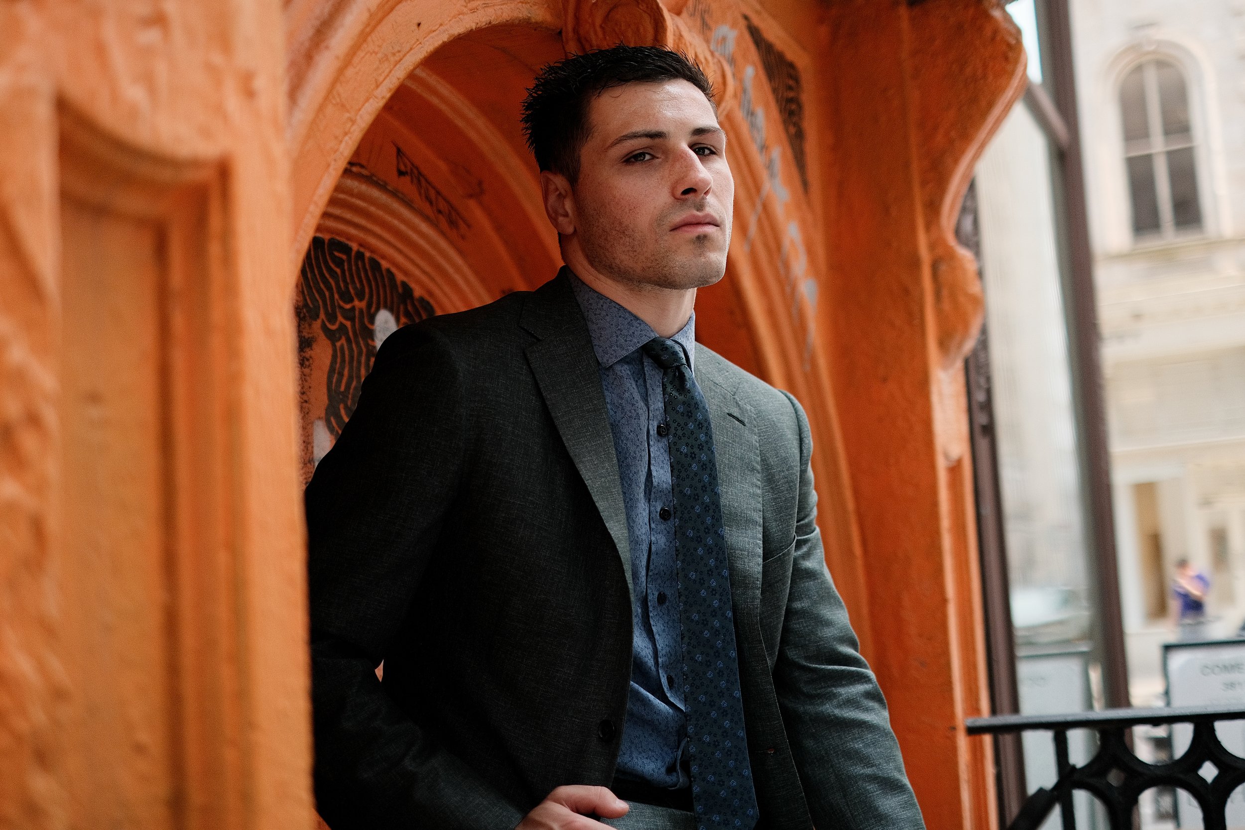 A young man with dark hair in a business suit standing near a window with orange architecture behind him