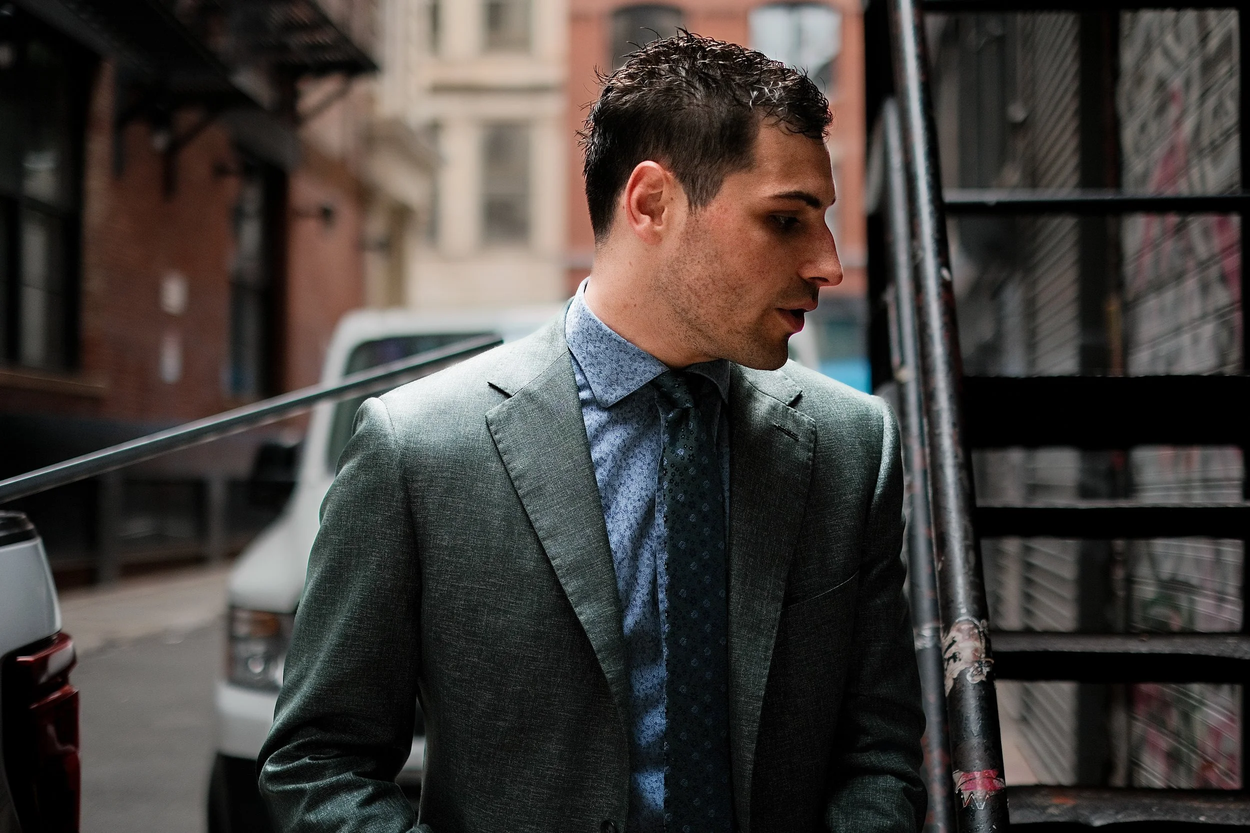 A man in a gray suit with a patterned dress shirt and tie standing outdoors near fire escape stairs in an urban alley, looking to the side.