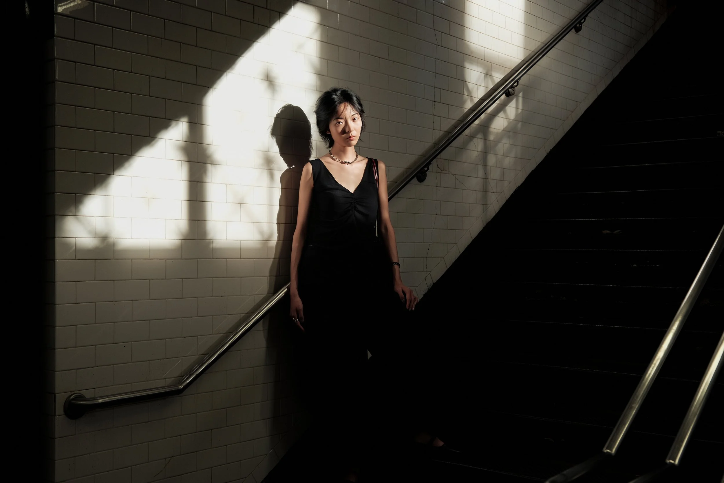 A woman with short black hair wearing a black sleeveless dress and necklace stands under a spotlight against a subway wall and on stairs, casting a shadow on the wall.