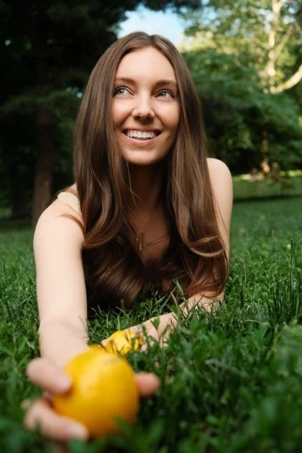 A young woman with long brown hair smiles while lying on grass in a park, holding a yellow fruit in her hand.