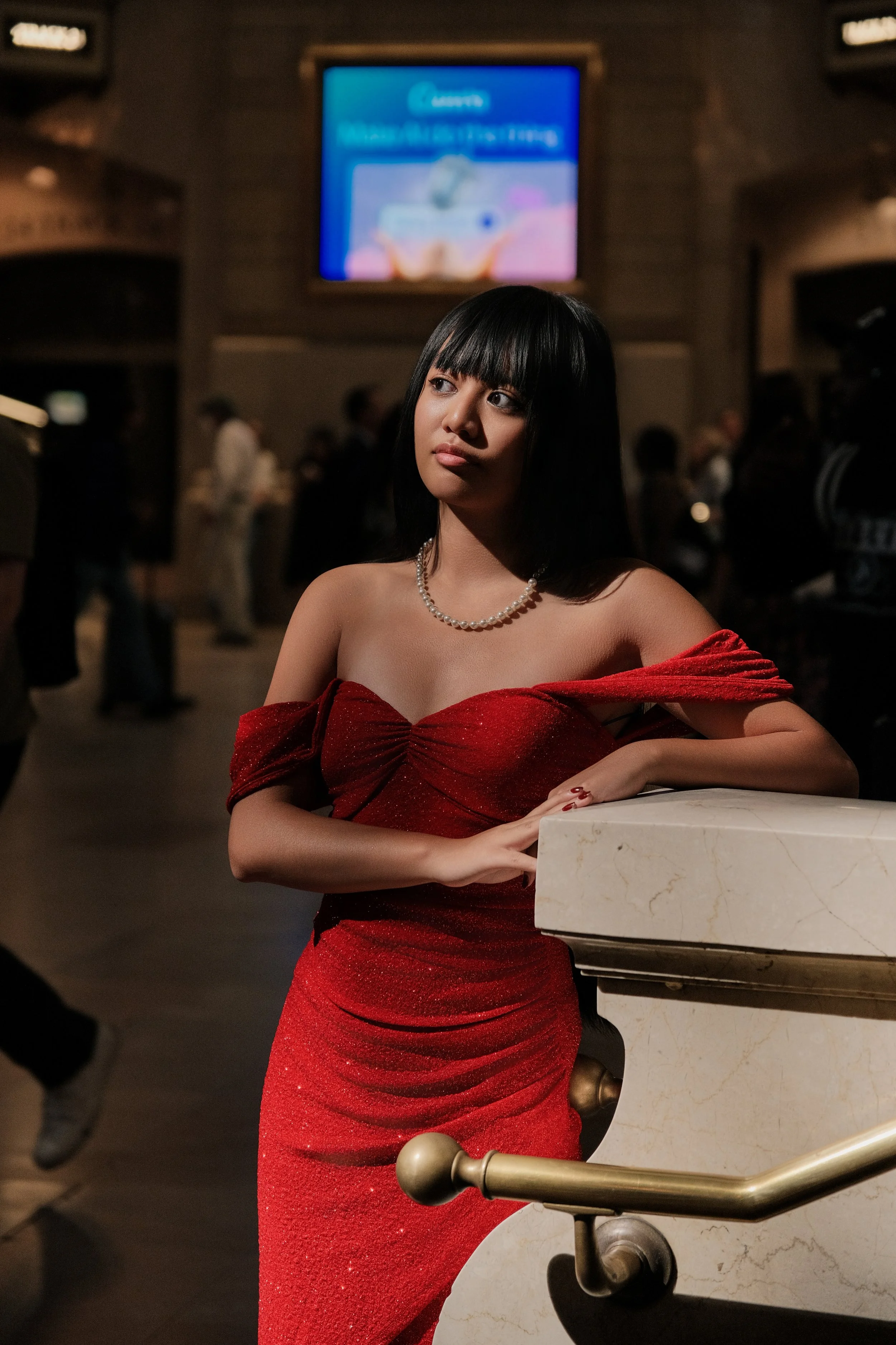 A woman with straight black hair and bangs, wearing a red off-shoulder dress and a pearl necklace, standing next to a marble railing inside a dimly lit venue with a large blue screen in the background.