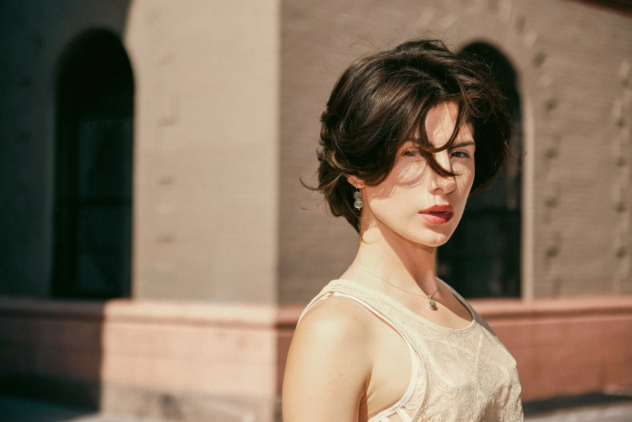 A woman with short, wavy dark hair and earrings is standing outdoors, wearing a light-colored sleeveless top, with sunlight highlighting her face and a blurred background.