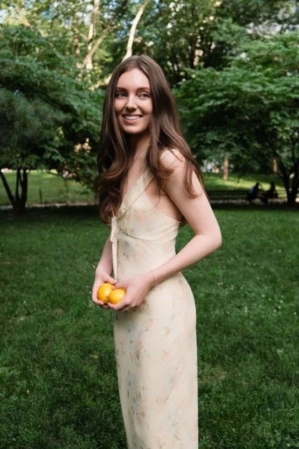 Young woman with long brown hair in a light-colored dress holding yellow fruit outdoors in a park with green grass and trees.