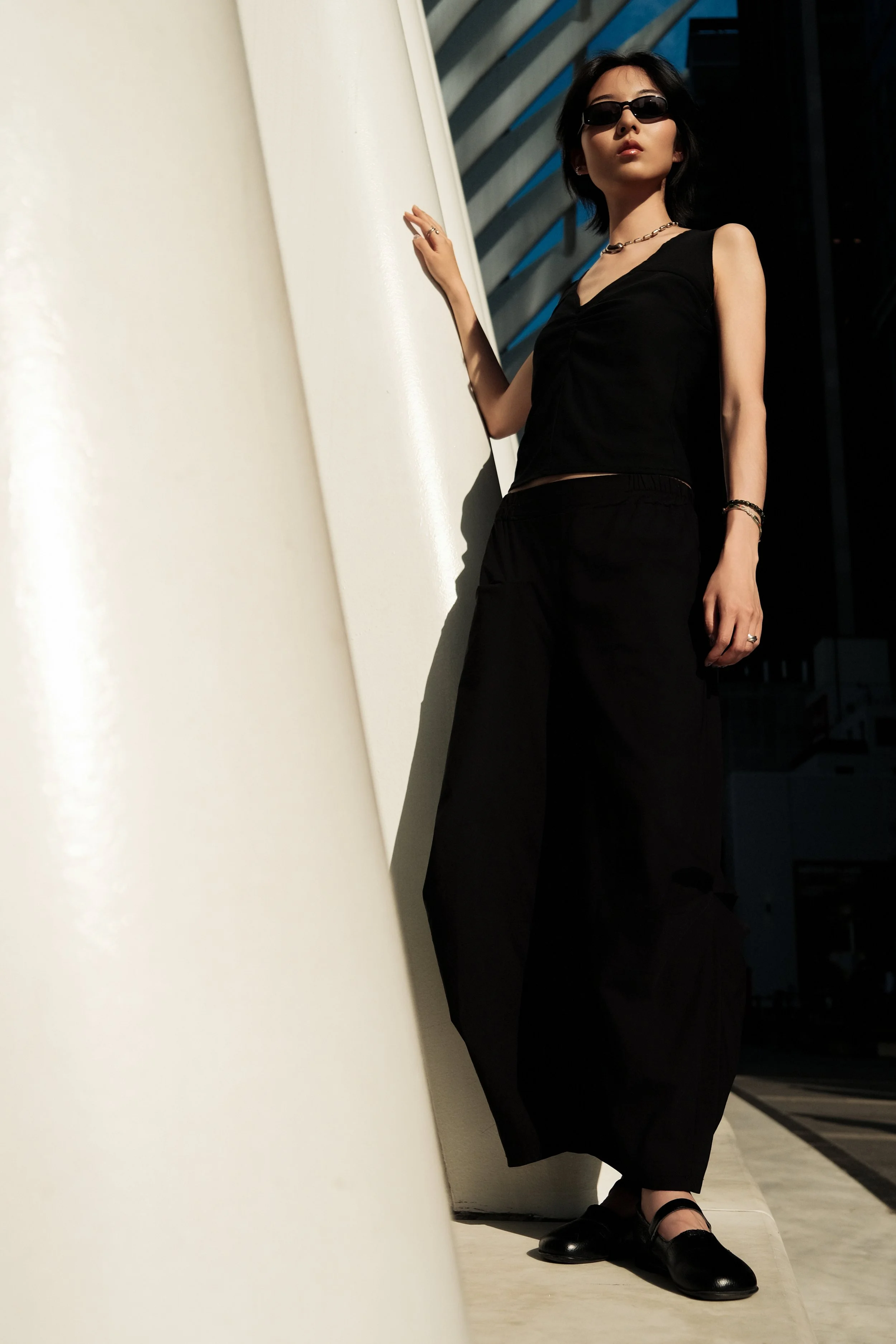 A woman with short dark hair, wearing sunglasses, a black sleeveless top, black pants, and black shoes, stands against a white wall in a modern building.