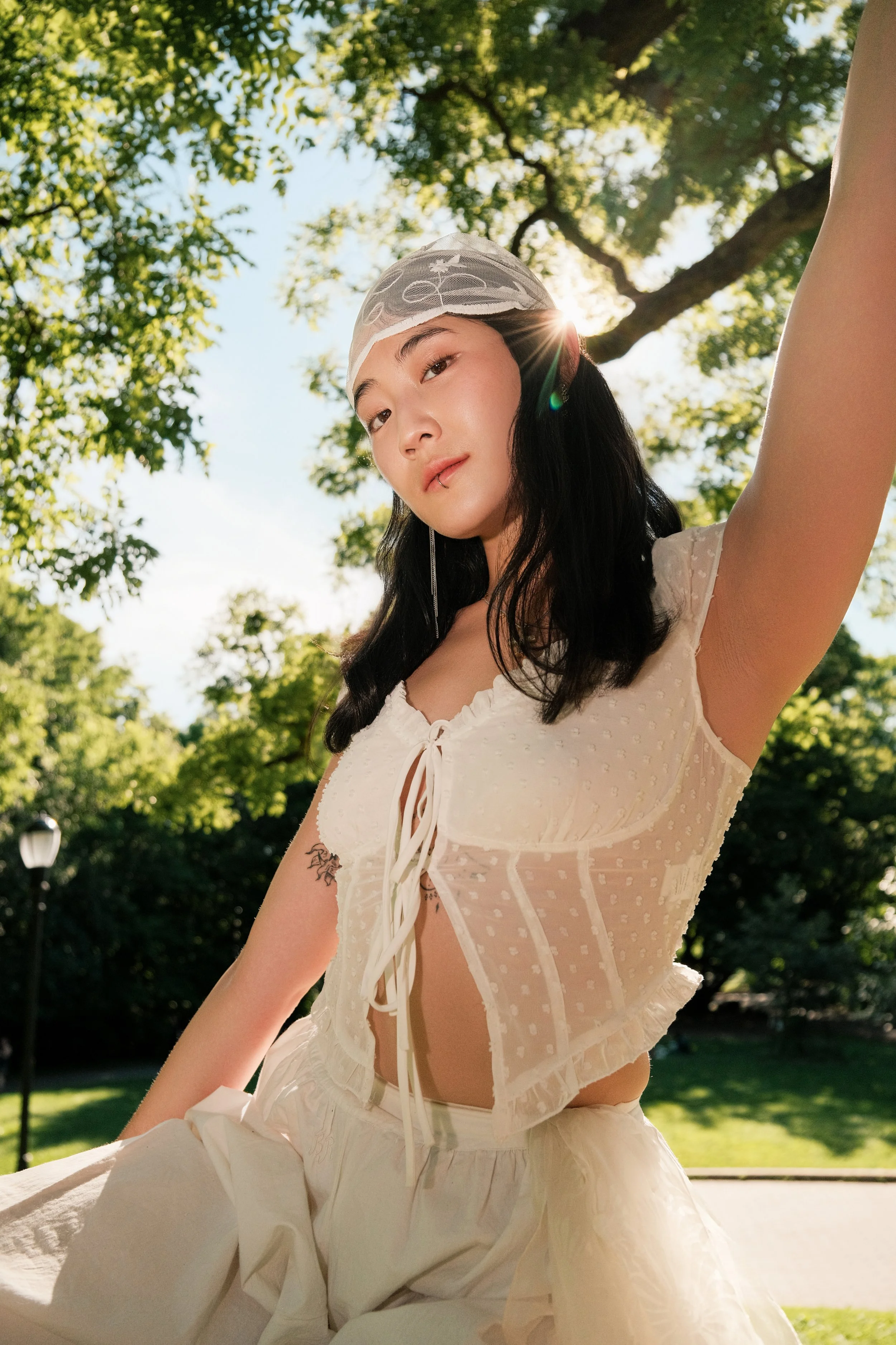 A young woman with long black hair wearing a white lace top and a sheer head covering poses outdoors in a green park, with sunlight filtering through the trees behind her.