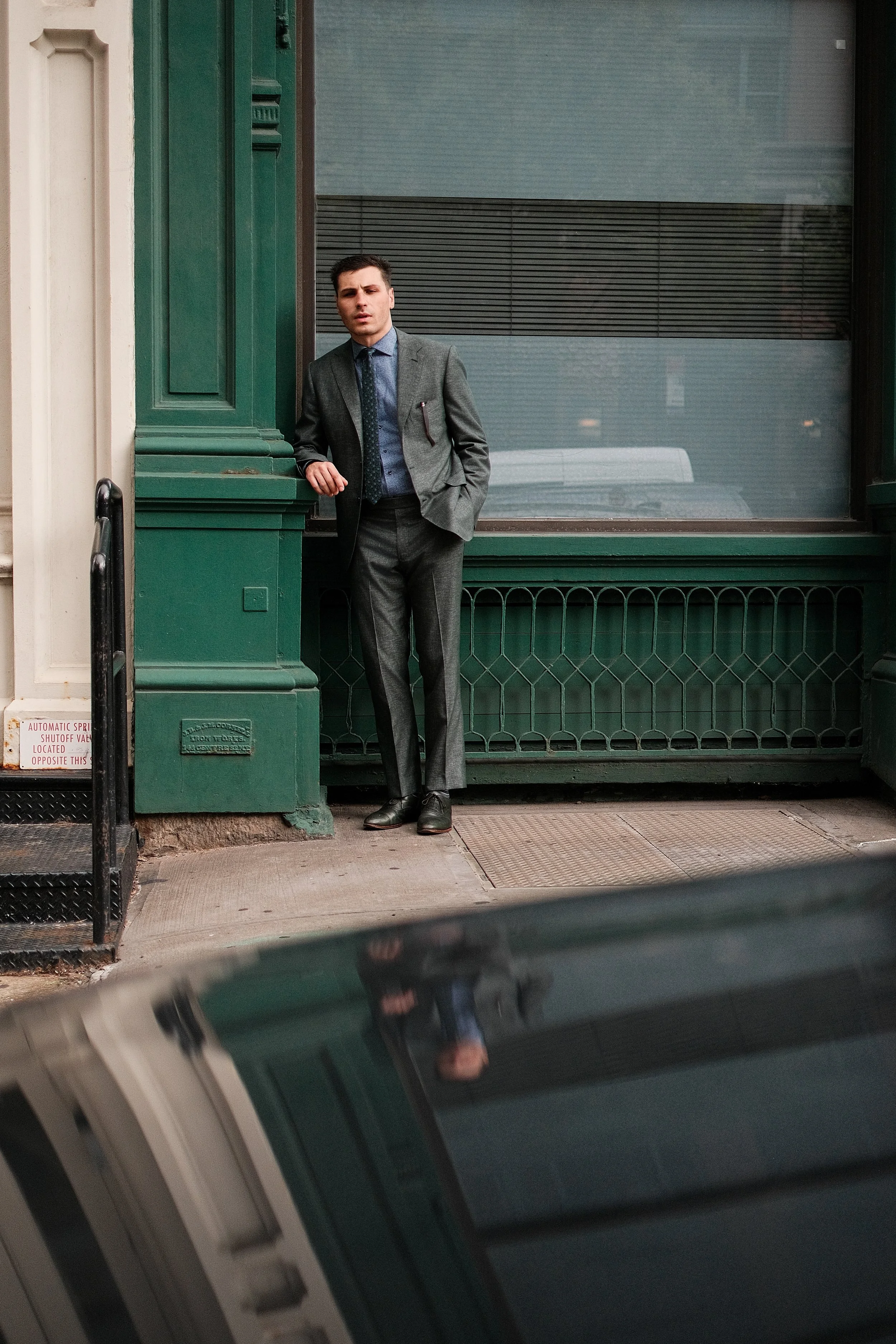 A man dressed in a gray suit with a blue shirt and tie, standing outside a building with green exterior walls and large windows, leaning against the wall with one hand in his pocket.