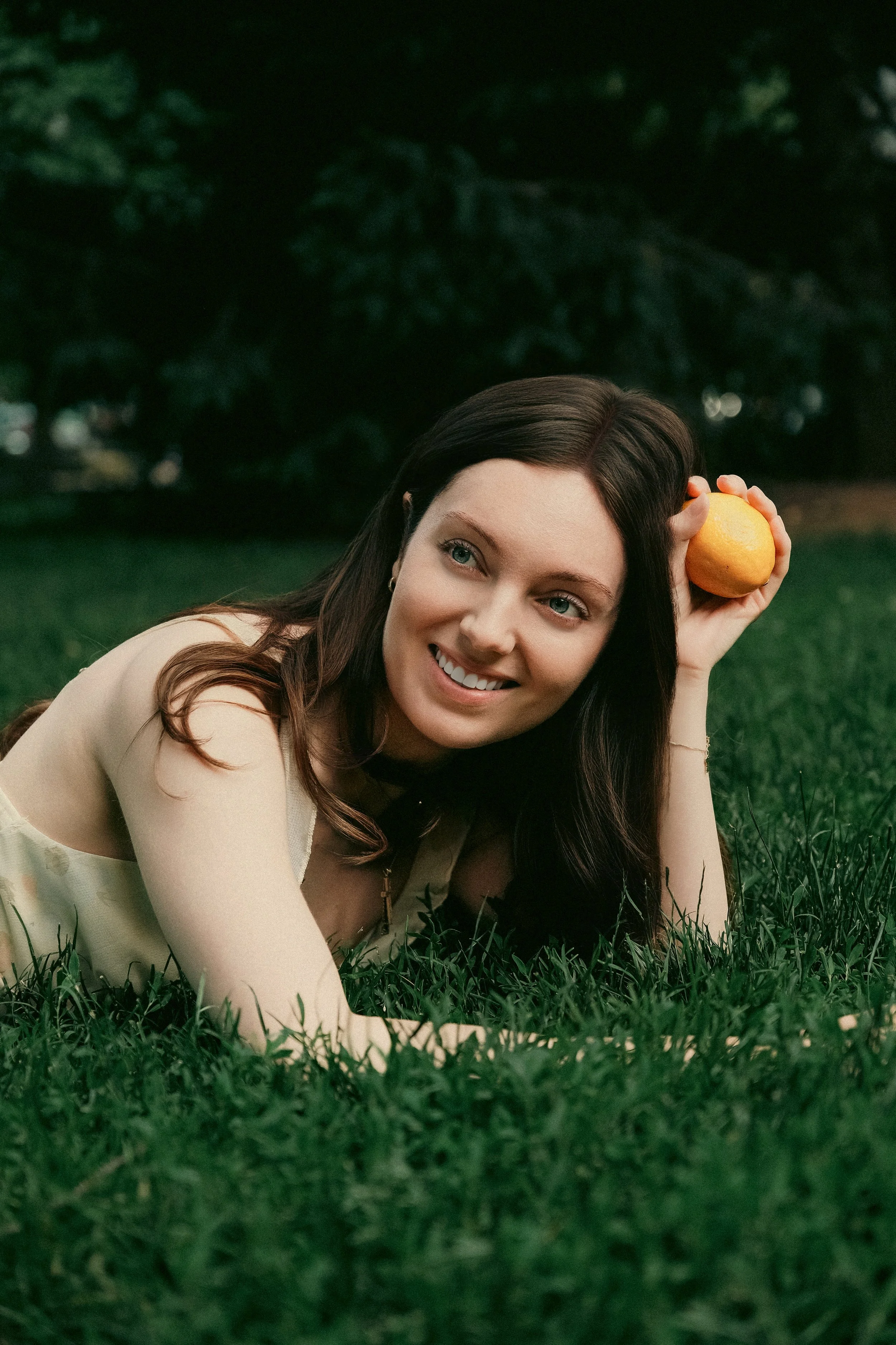 A woman with long brown hair and blue eyes lying on green grass, holding a yellow lemon to her head, smiling, with dark green trees in the background.