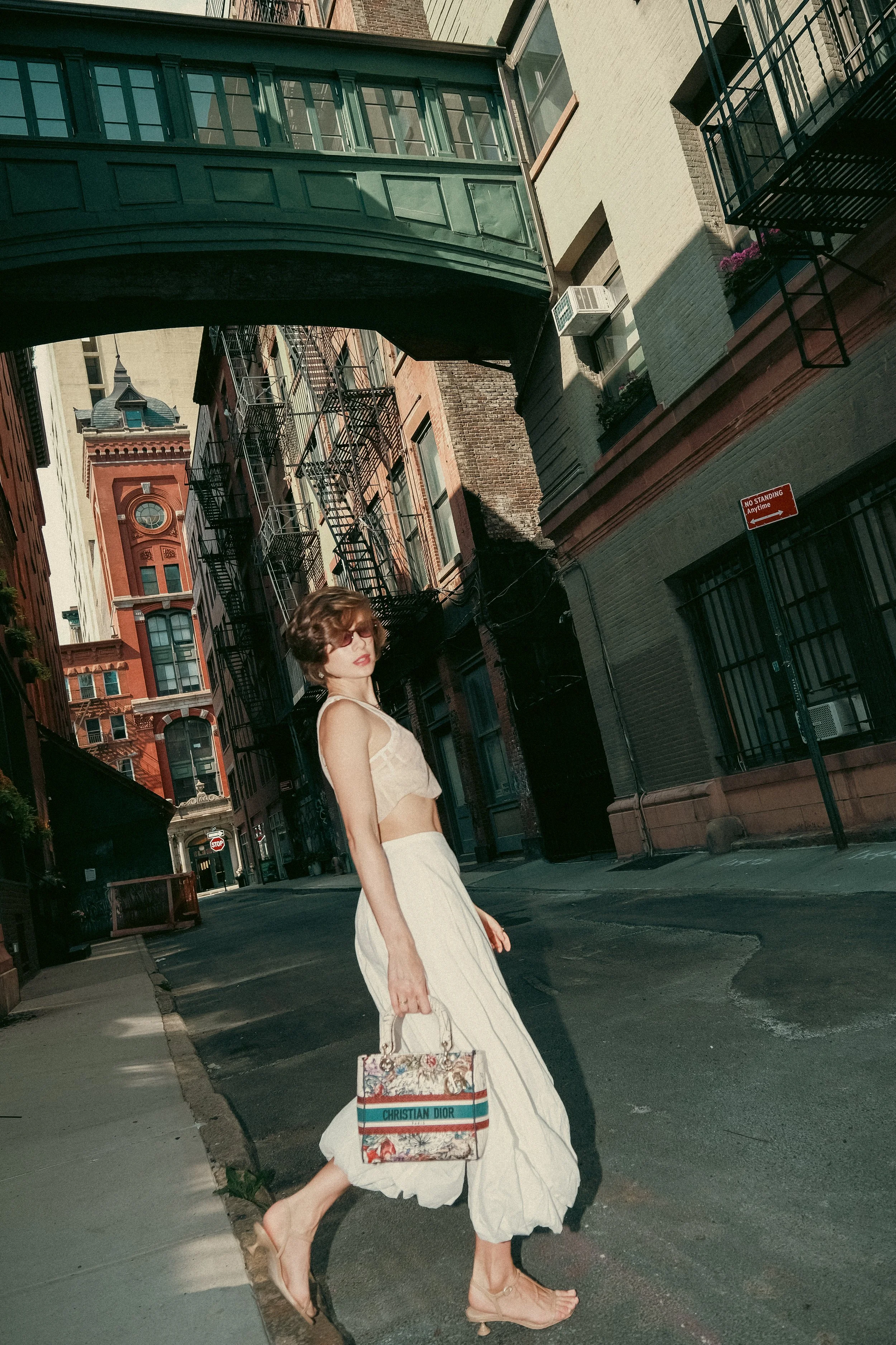 A woman with short, wavy hair and sunglasses is standing on a city street, holding a Christian Dior handbag. She is wearing a cream-colored sleeveless top with a low back and a flowing white skirt. She is also wearing transparent heeled sandals. The 