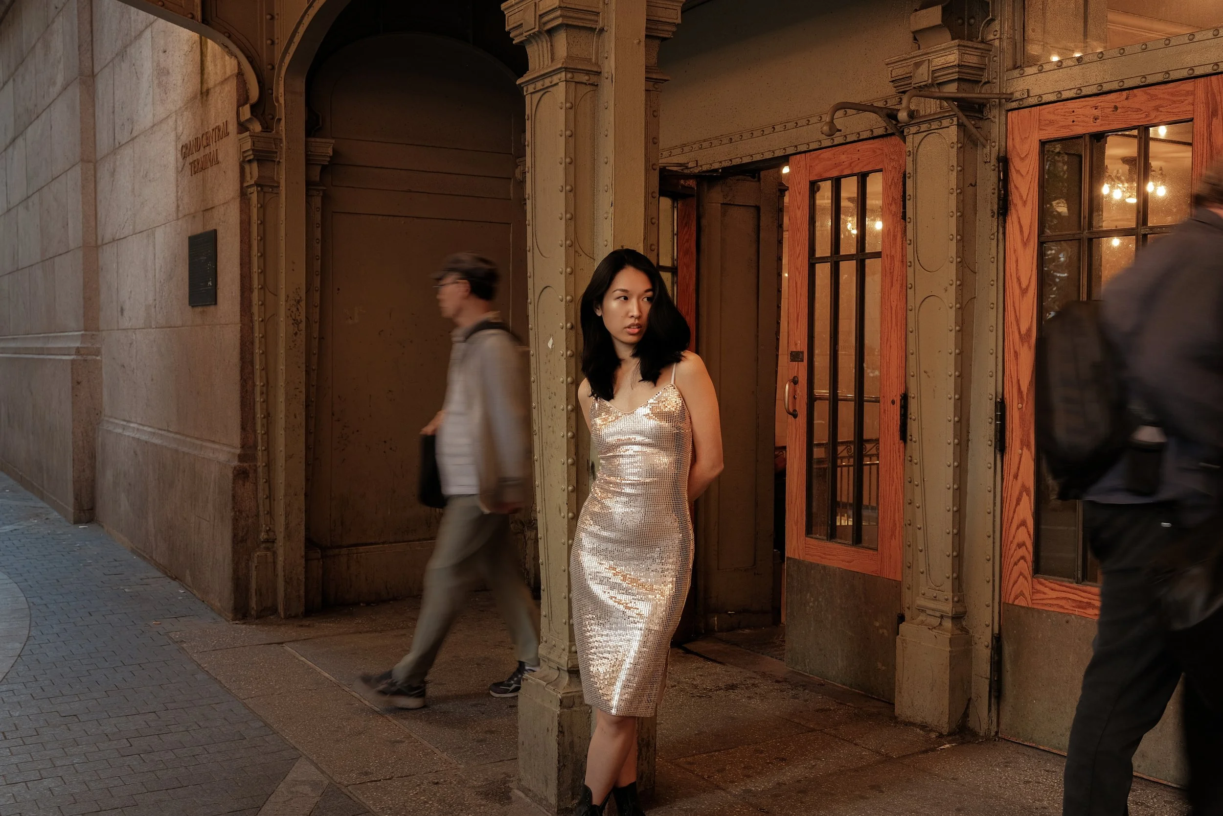 A young woman in a shimmering, silver, sequined dress standing near a corner outside a building with large wooden window frames. Two men walk by, one blurred in motion and the other partially visible.