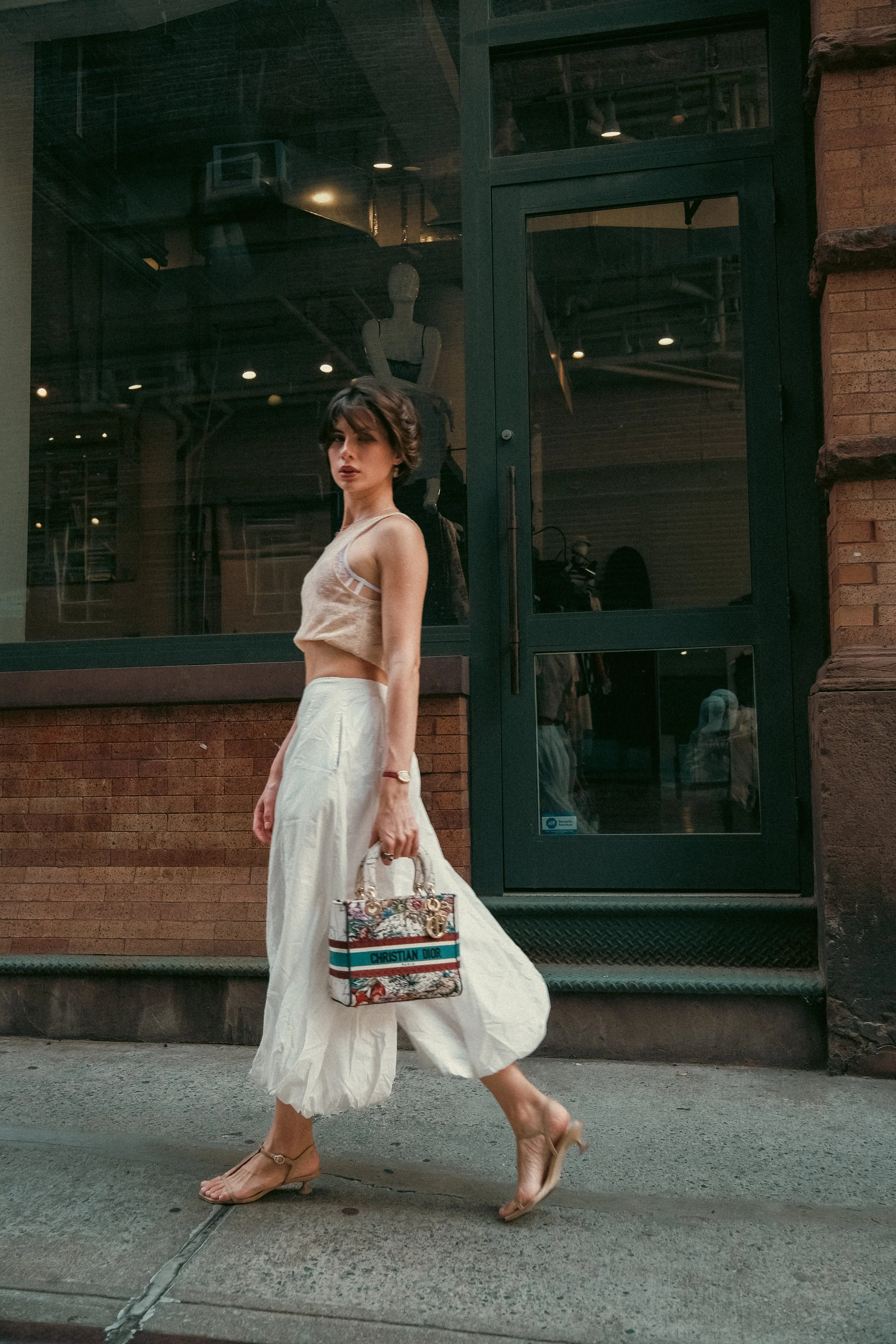 A woman in a beige lace crop top, white wide-leg pants, and beige heels, walking on the sidewalk while carrying a Christian Dior handbag. She has short dark hair and is looking at the camera. The background shows a building with a large glass window 
