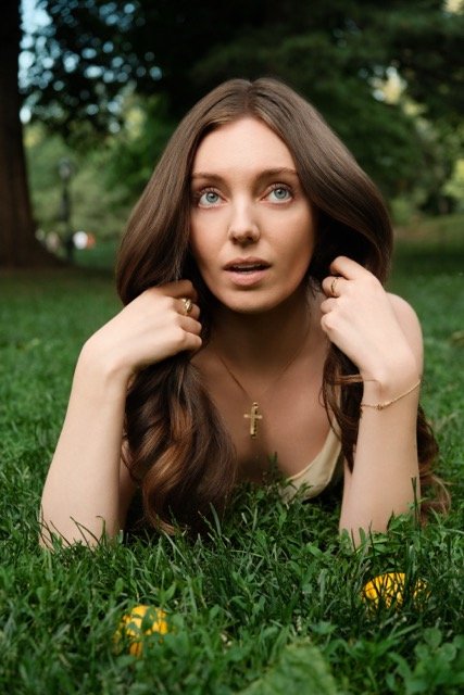 A young woman lying on grass outdoors with trees in the background, wearing a gold cross necklace and a cream-colored top, gazing at the camera.