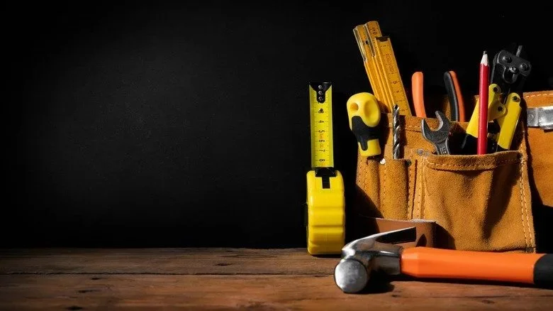 Tools in a pouch on a wooden surface with a dark background, including tape measure, hammer, screwdriver, tape, ruler, and other tools.