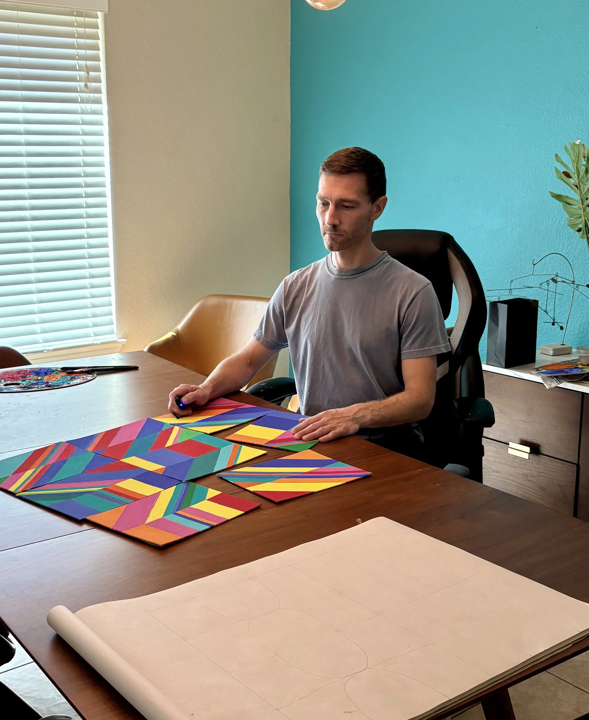 A man sitting at a table working on a geometric art project with painted canvas boards on a large table inside a room with teal and beige walls, blinds on the window, and a potted plant nearby.