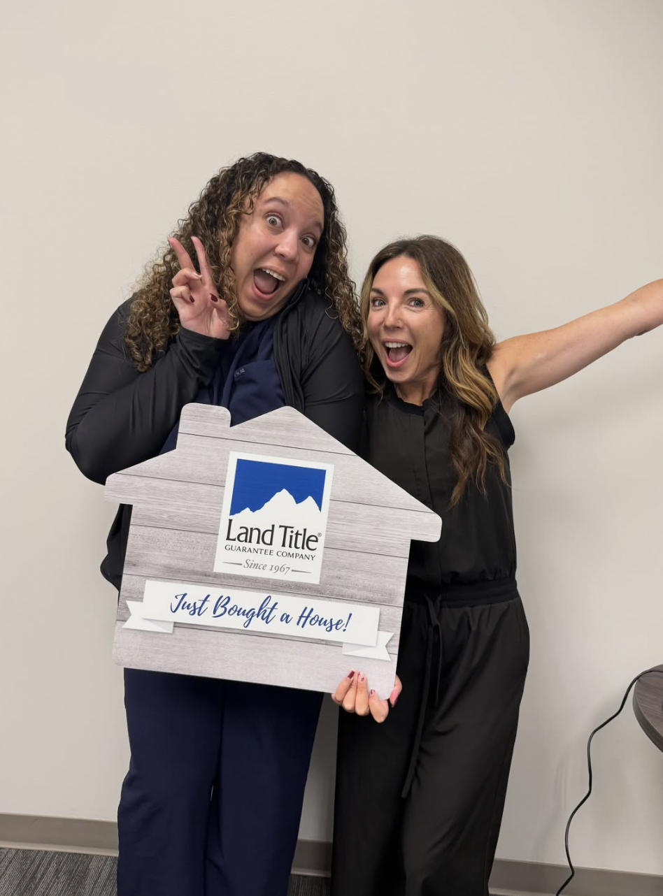 Two women celebrating with a house-shaped sign from Land Title, announcing they just bought a house, standing against a plain wall. South Denver Realtor serving Highlands Ranch, Lone Tree and Littleton.