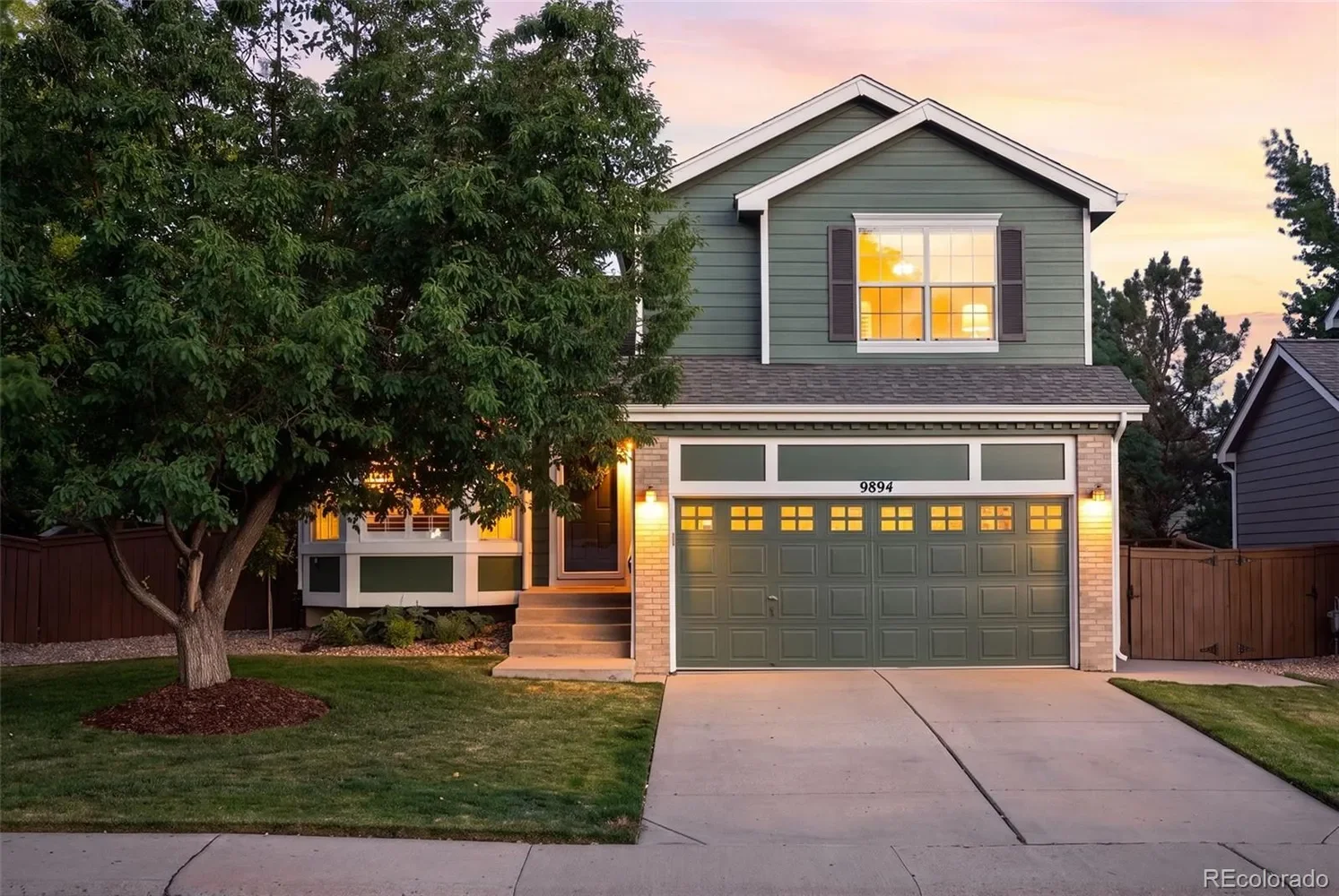 A two-story house with a green exterior and a two-car garage, illuminated by exterior lights at sunset, featuring a front lawn and a large tree on the left side.