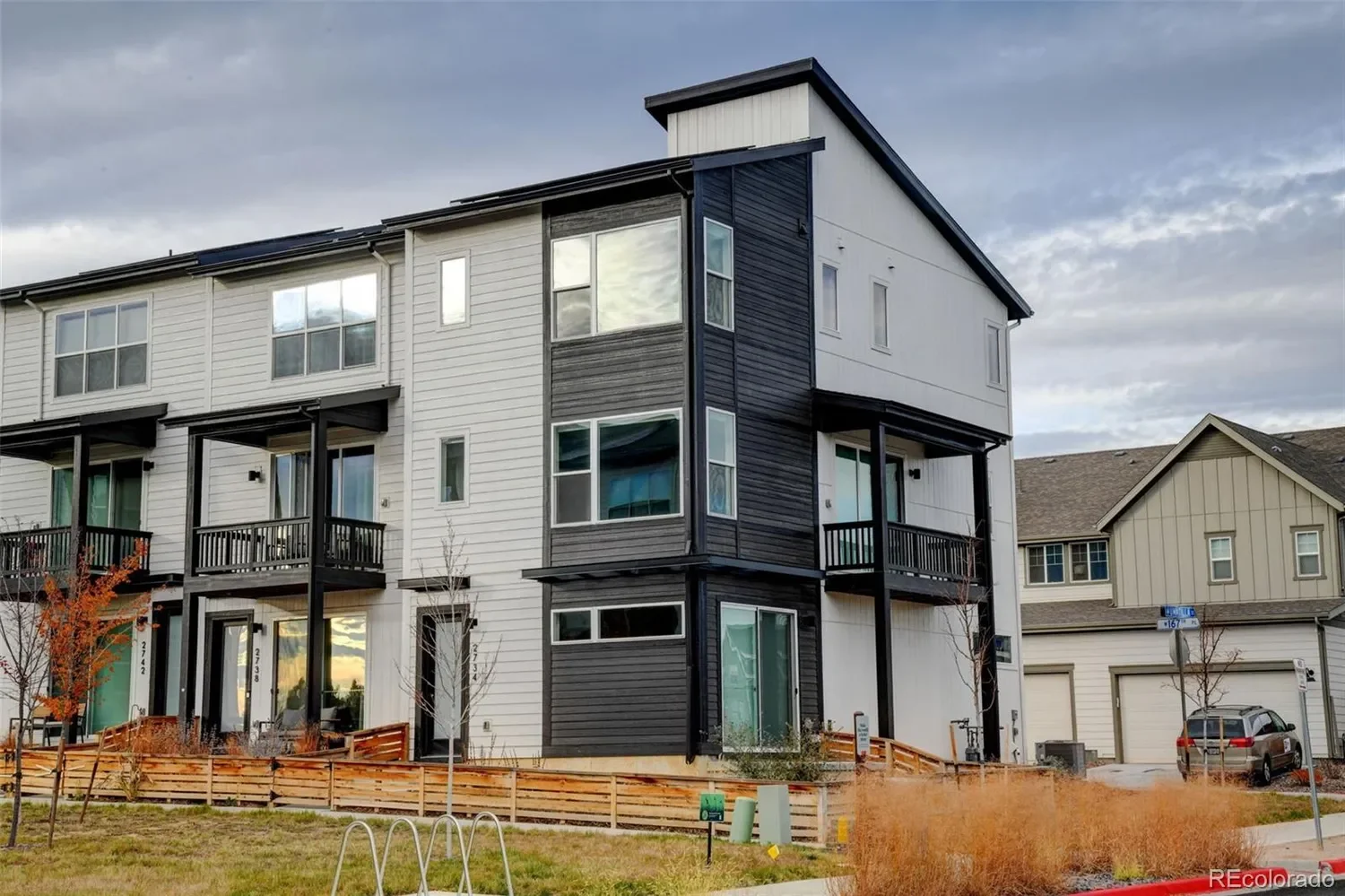 Modern three-story residential building with white and black exterior, multiple large windows, small balconies, and a gated front yard with young trees and a grassy area.